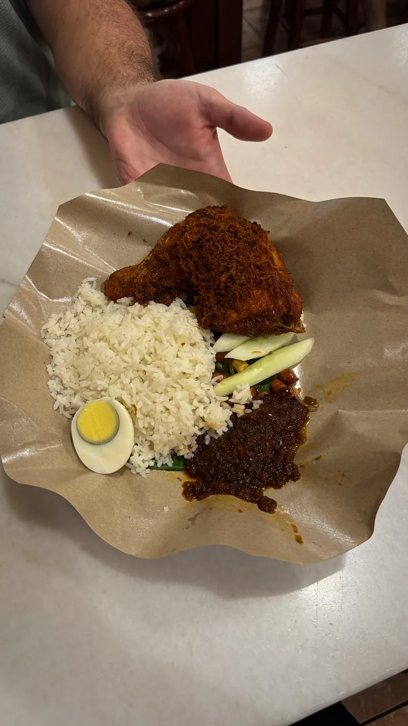 A Village Park nasi lemak held up on brown paper, with the fried chicken thigh covered in crispy bits