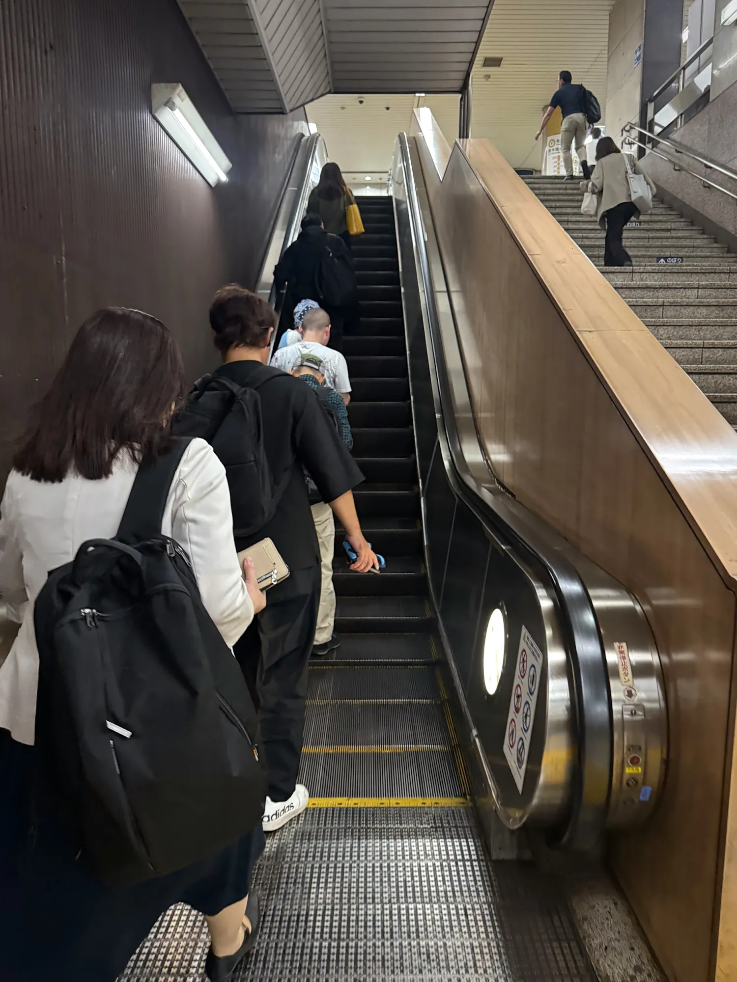 Commuters with backpacks riding single file up a long subway escalator, with a staircase running alongside.