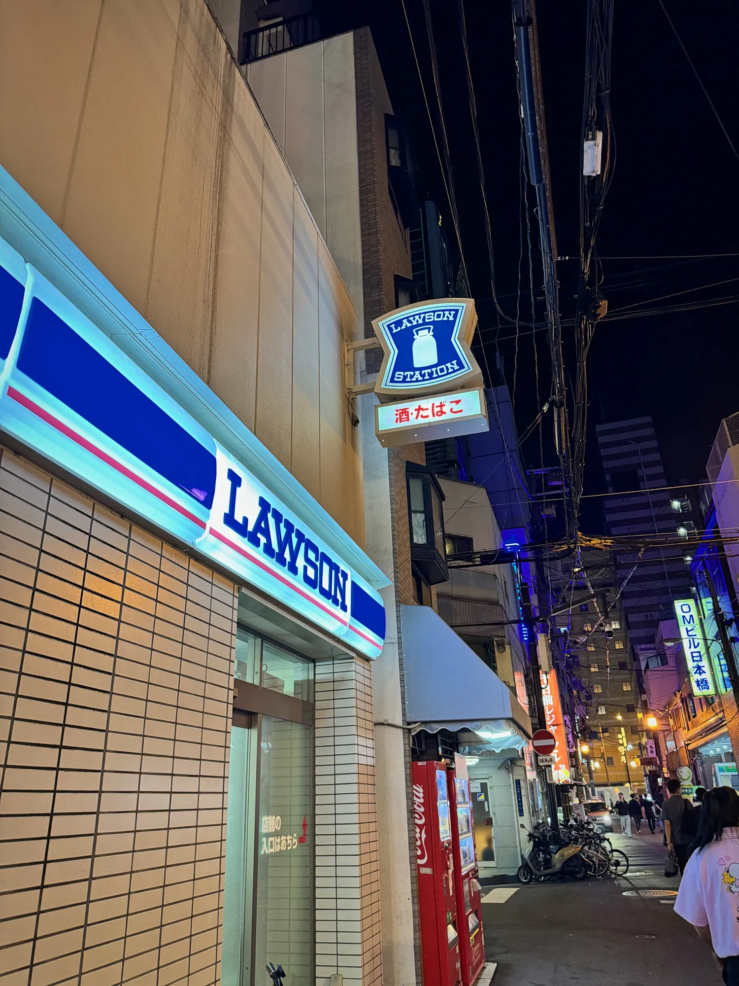 A Lawson convenience store lit up blue and white at night on a narrow Osaka street.