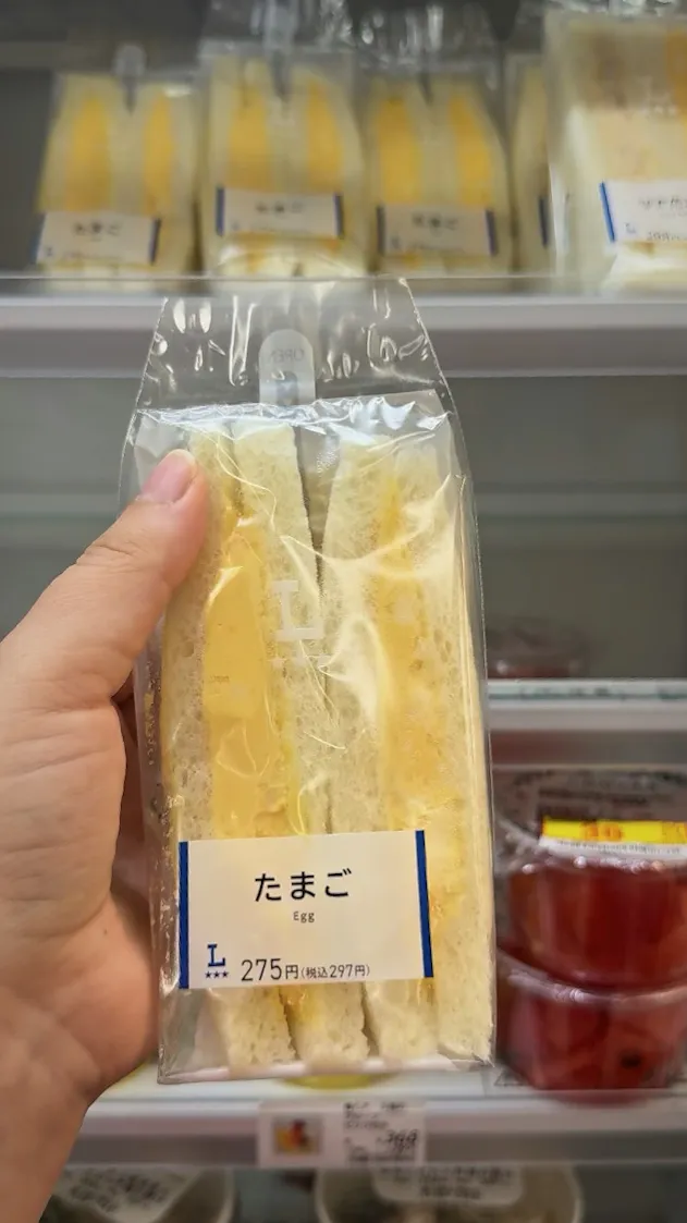 A hand holding a Lawson egg sandwich priced at 275 yen in front of the sandwich shelf in Osaka.