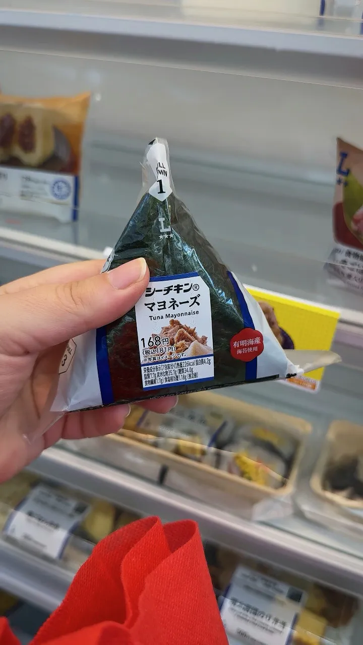 A hand holding a Lawson Sea Chicken tuna mayonnaise onigiri in front of a refrigerated shelf in Osaka.
