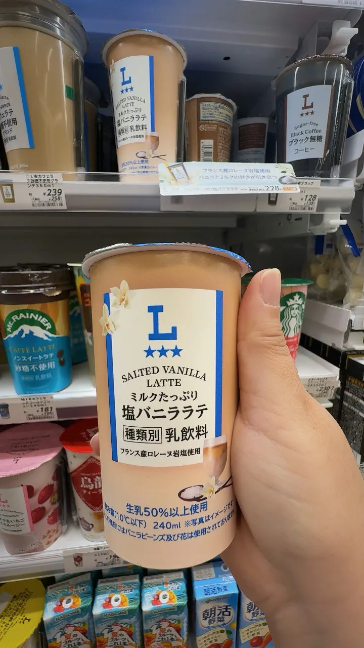 A hand holding a Lawson salted vanilla latte cup in front of the chilled drinks shelf in Osaka.