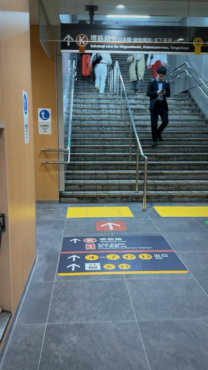 A traveler pulling a red suitcase down a long subway staircase under signs for the Sakaisuji Line in Osaka, with floor arrows pointing to the exits.