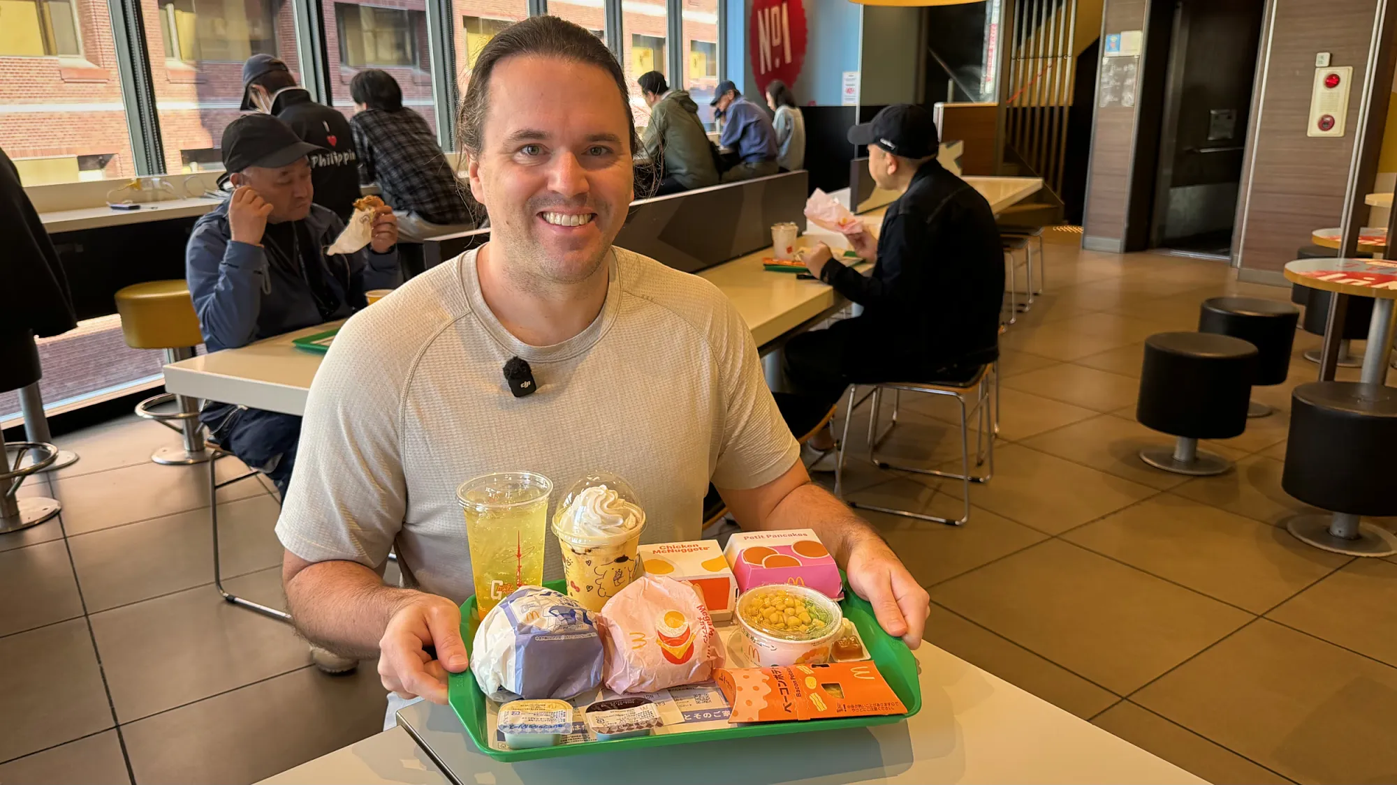 Cream smiling inside a McDonald's in Shinjuku, Tokyo, holding a green tray with a Chicken Tatsuta, a Mega Muffin, Petite Pancakes, Chicken McNuggets, corn and edamame, a Bacon Potato Pie, and two drinks.