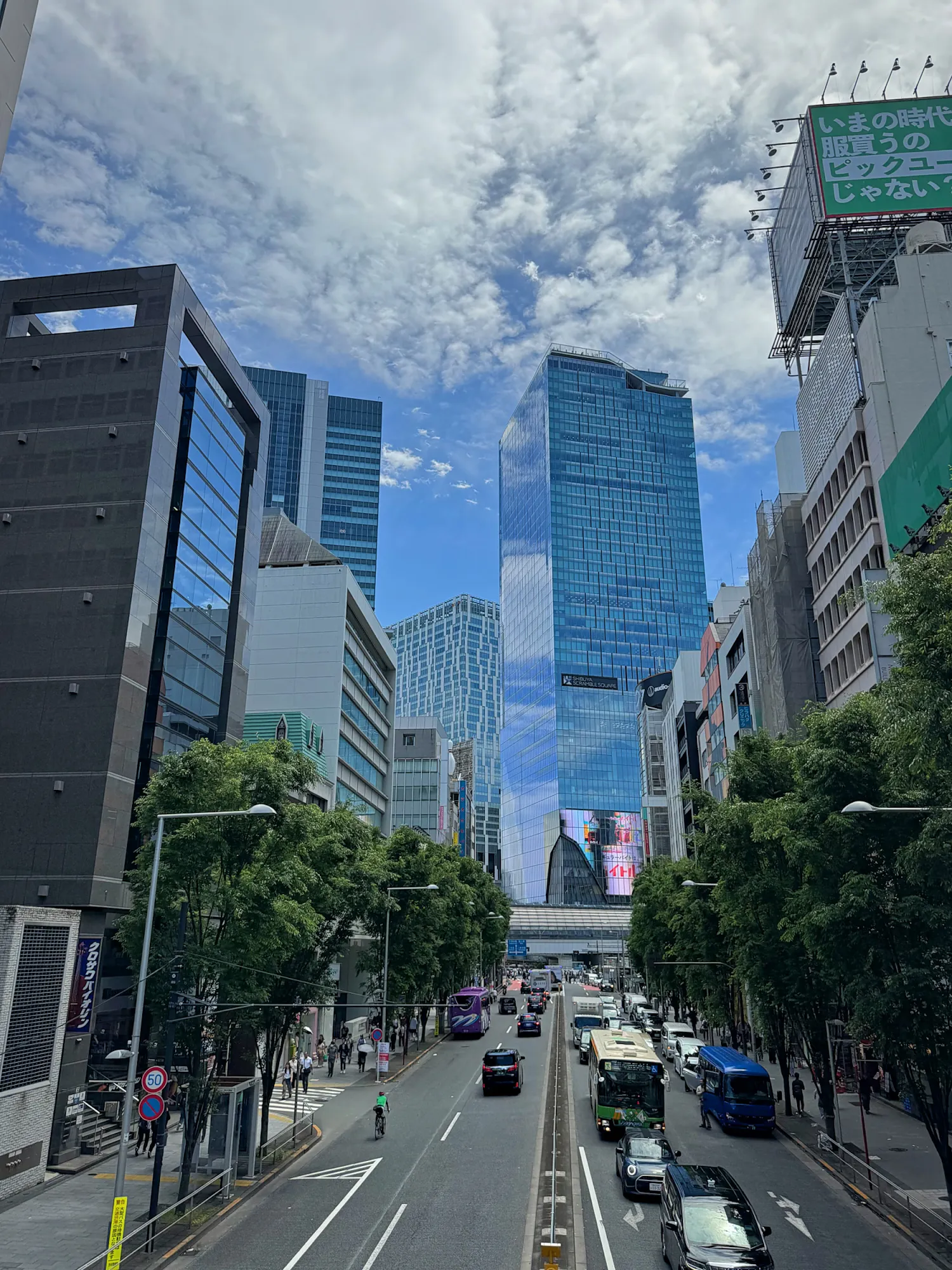 A wide, tree-lined avenue running between glass skyscrapers in Tokyo, Japan, under a blue sky with scattered clouds.