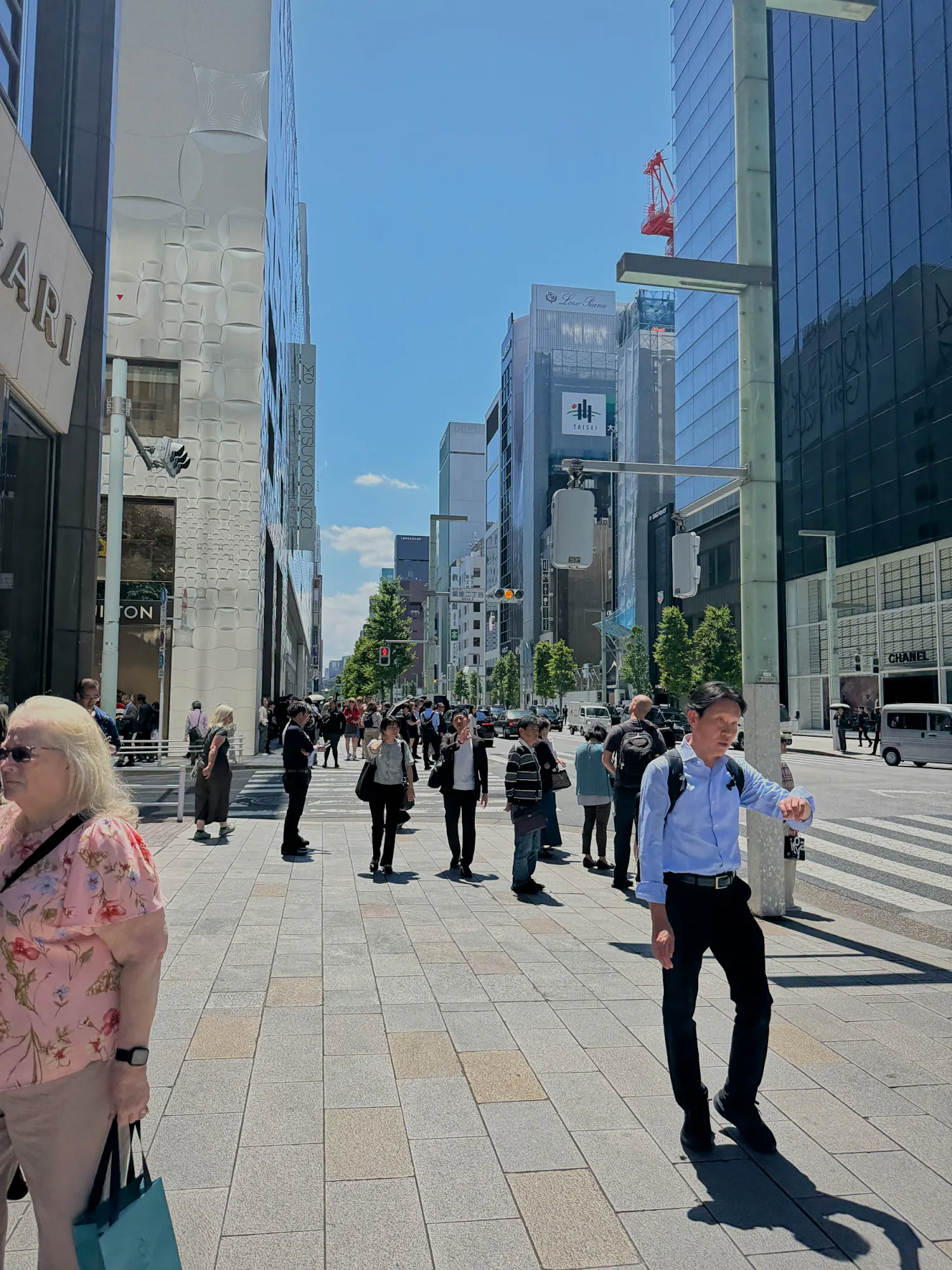Shoppers walking along a wide, sunny Ginza street lined with glass department stores in Tokyo.