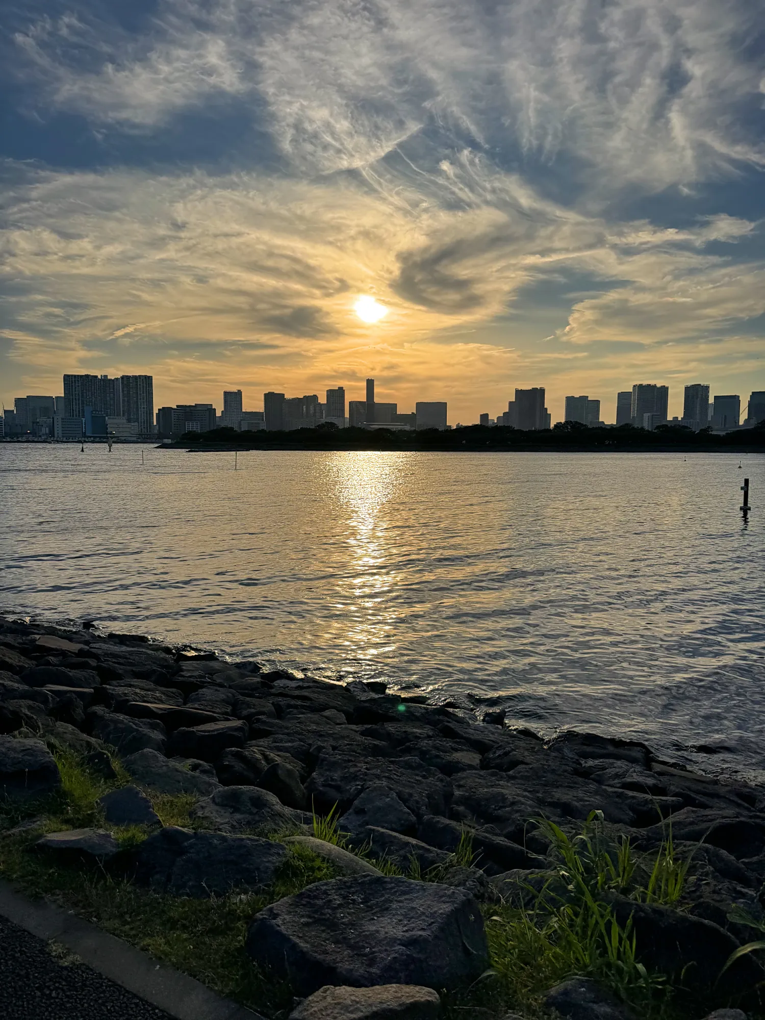 The Tokyo skyline silhouetted against an orange sunset over the water of Tokyo Bay, with rocks along the shore.