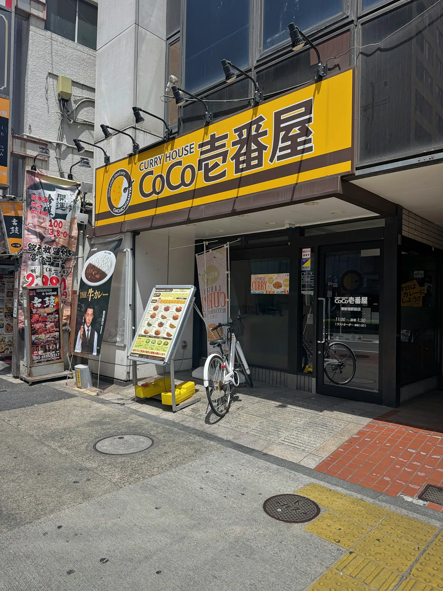 The yellow Curry House CoCo Ichibanya sign above the glass entrance of the Higashi-Shinjuku Ekimae branch in Tokyo, with a menu board and a parked bicycle out front.