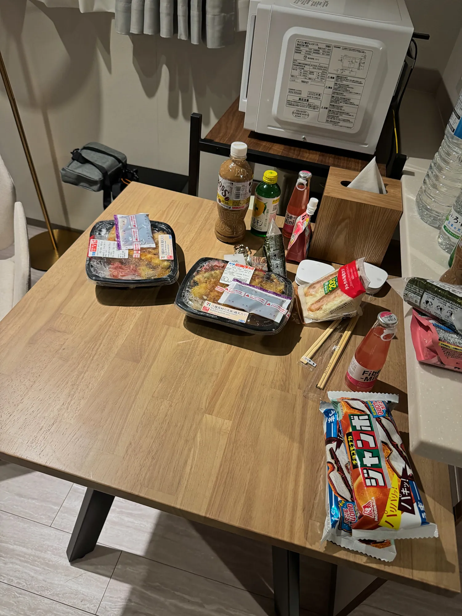 A wooden table in a small Tokyo apartment covered with convenience store bento boxes, snacks, and drinks.