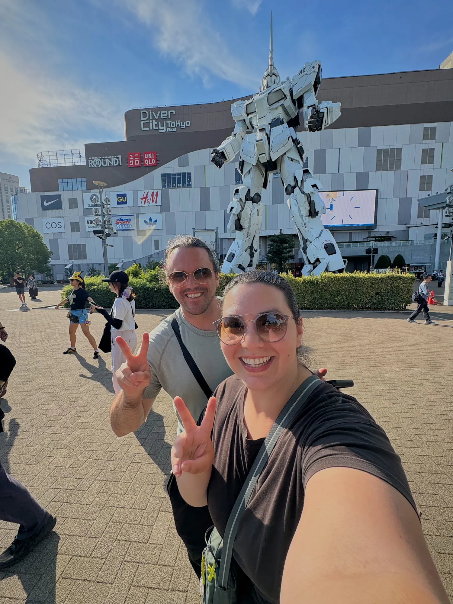 Bernie and Cream smiling and making peace signs in a selfie in front of the giant Unicorn Gundam statue in Odaiba, Tokyo.