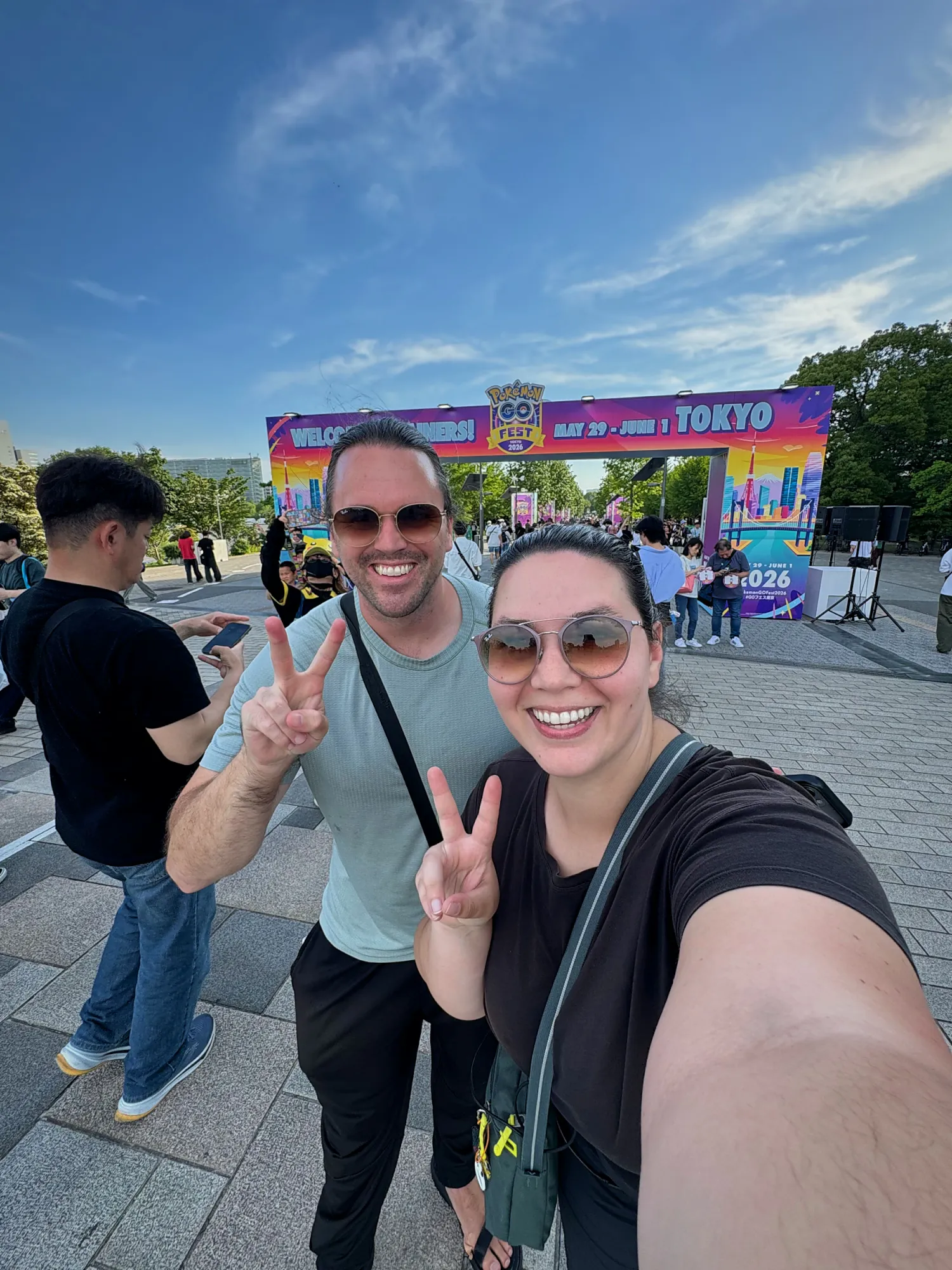 Bernie and Cream taking a selfie with peace signs in front of the colorful Pokémon GO Fest Tokyo 2026 entrance arch by Tokyo Bay.