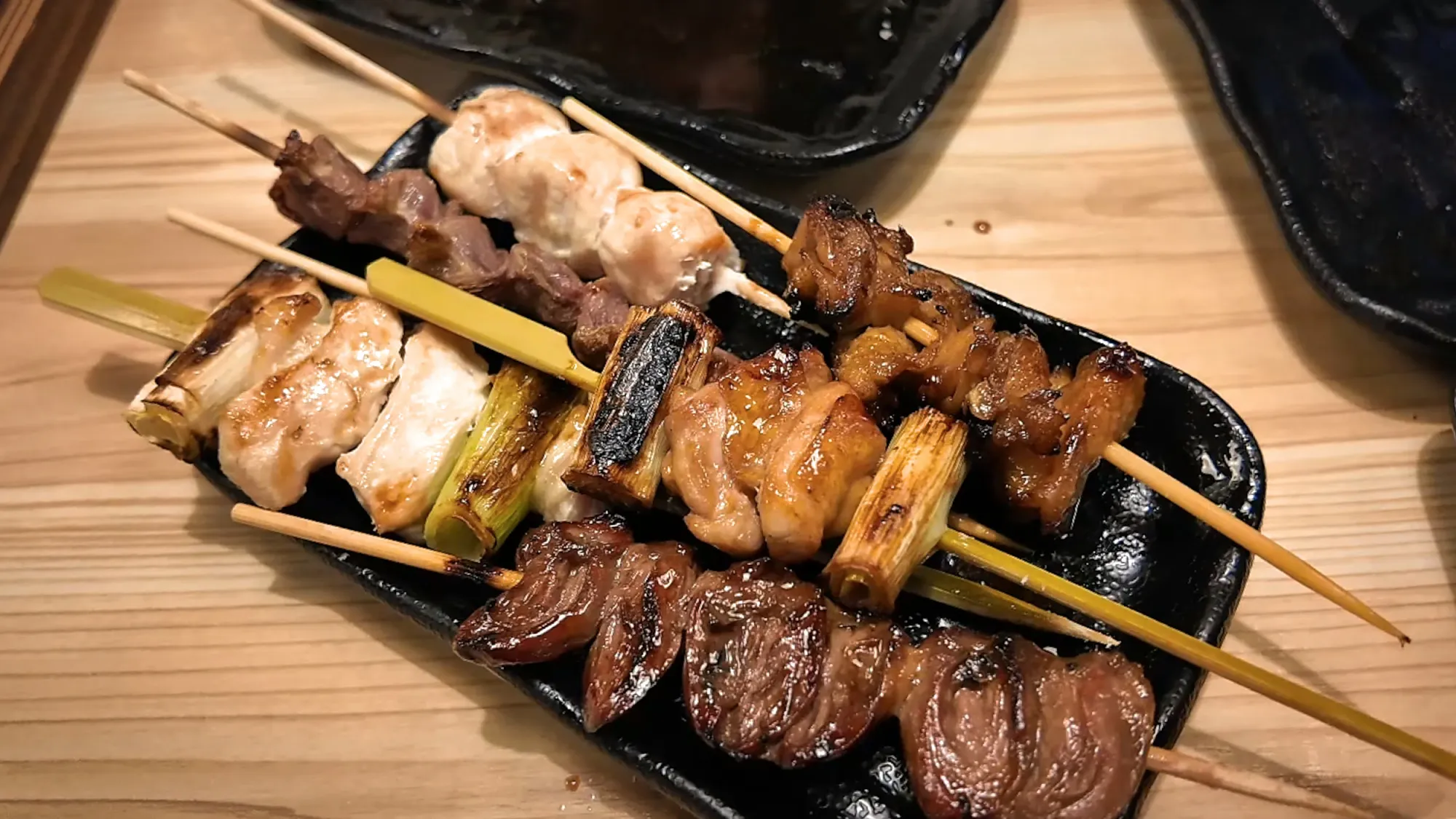A black plate of assorted yakitori skewers, including salted chicken breast with leek, sauced thigh, and hearts, at Torikizoku in Tokyo.