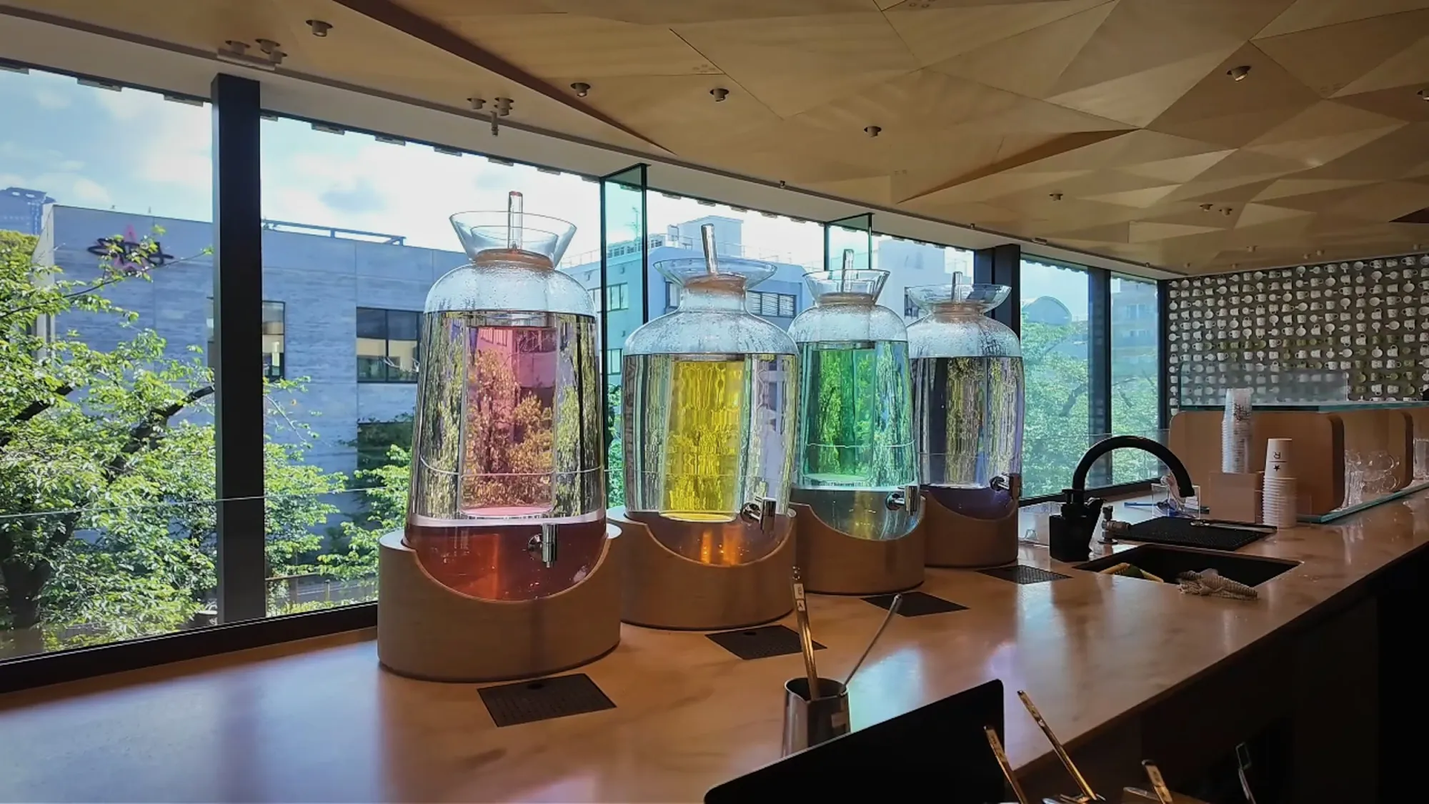 Large glass dispensers of red, yellow, and green iced tea on a wooden counter by the windows at Starbucks Reserve Roastery Tokyo.