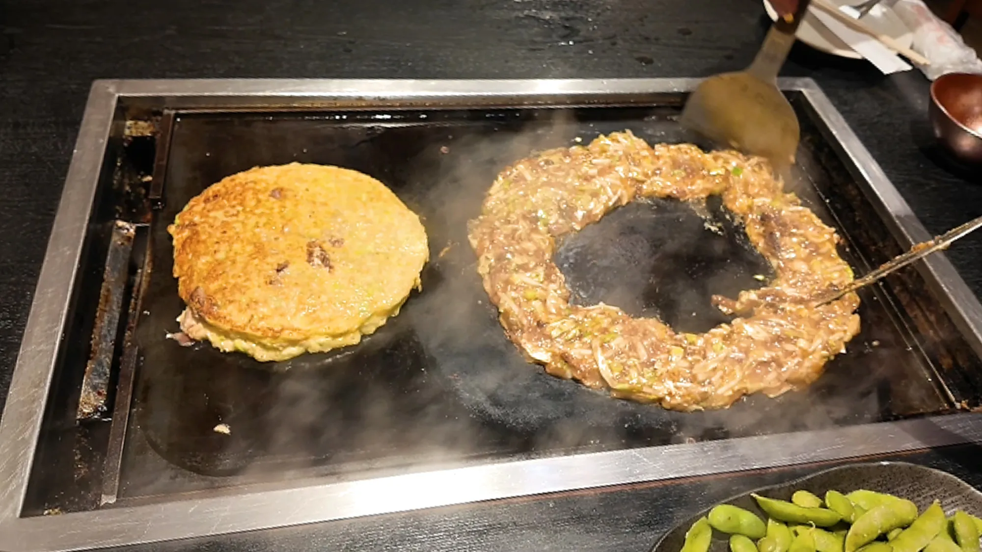 A round okonomiyaki pancake beside a ring of beef and cabbage monjayaki cooking on a teppan griddle at Osakaya, Tokyo.