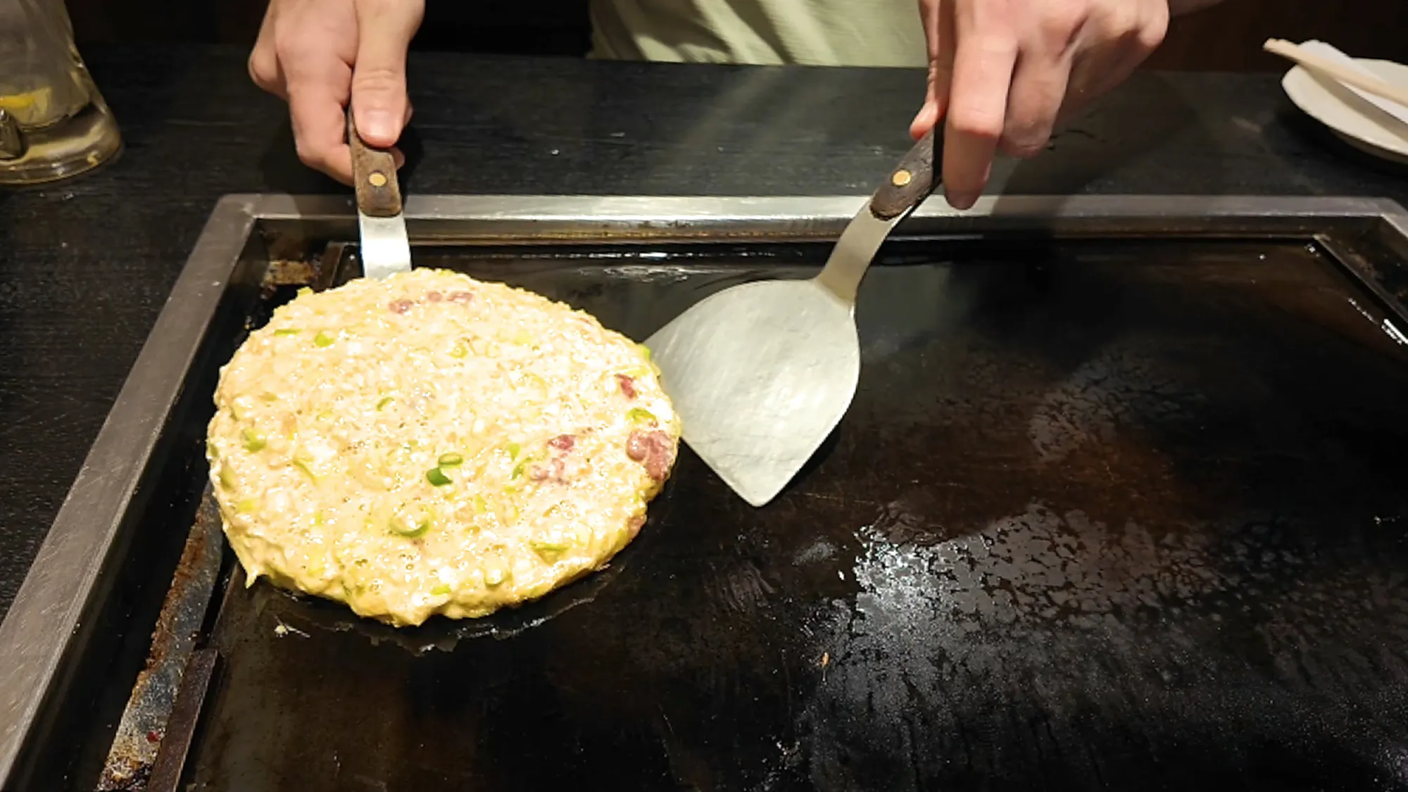 Two metal spatulas shaping a round okonomiyaki pancake full of cabbage and green onion on a hot teppan griddle at Osakaya, Tokyo.