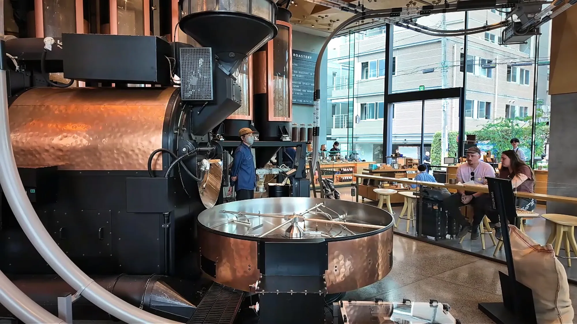 A large copper coffee roaster on the first floor of Starbucks Reserve Roastery Tokyo, with tables and big windows behind it.