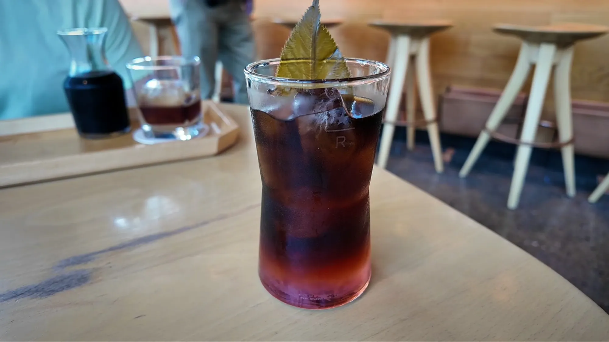 A tall glass of the Sakura barrel-aged cold brew over ice with a leaf garnish on a wooden table at Starbucks Reserve Roastery Tokyo.