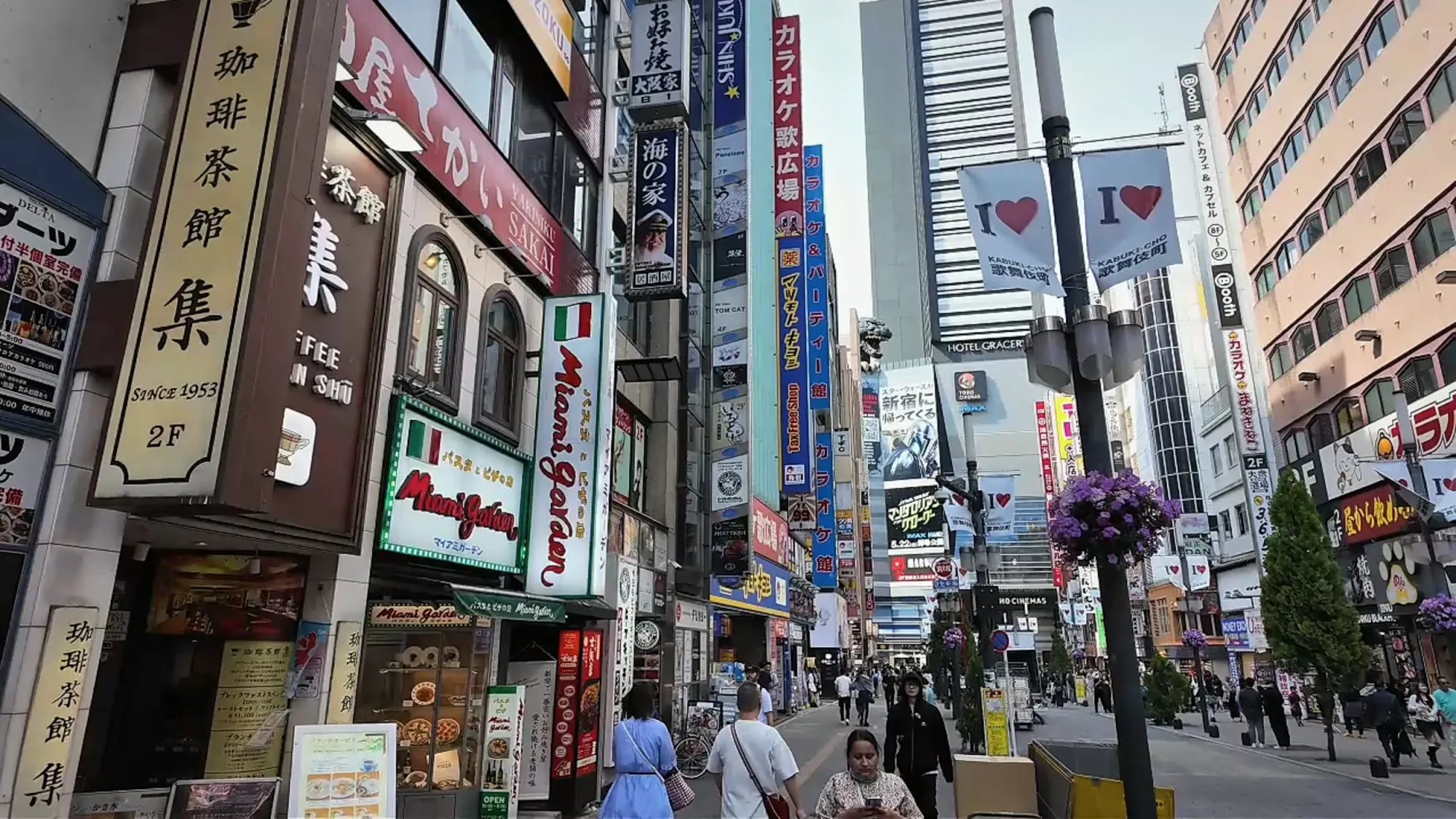 A sunny Kabukicho street in Shinjuku, Tokyo, lined with shop signs, billboards, and tall buildings.