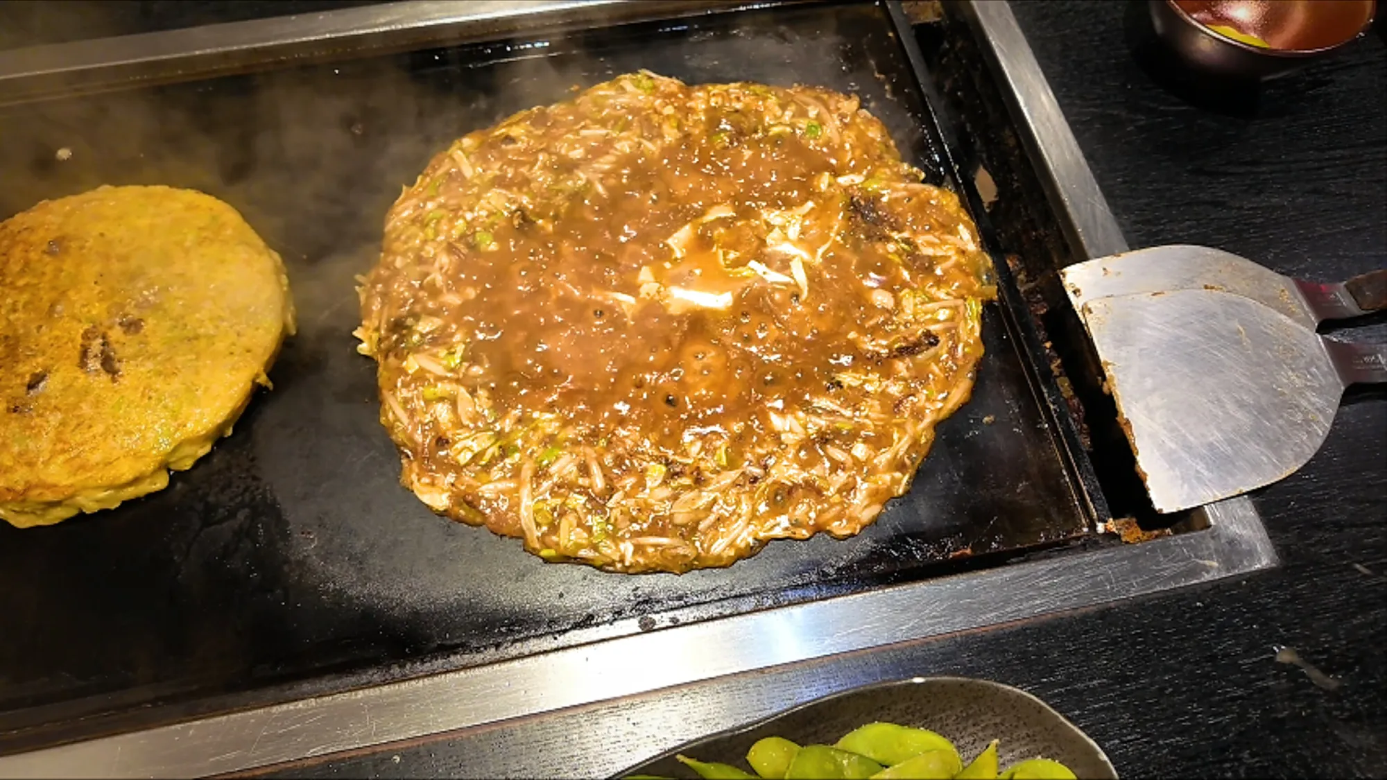Beef monjayaki spread thin and bubbling into a gooey, caramelized layer on a teppan griddle beside an okonomiyaki at Osakaya, Tokyo.