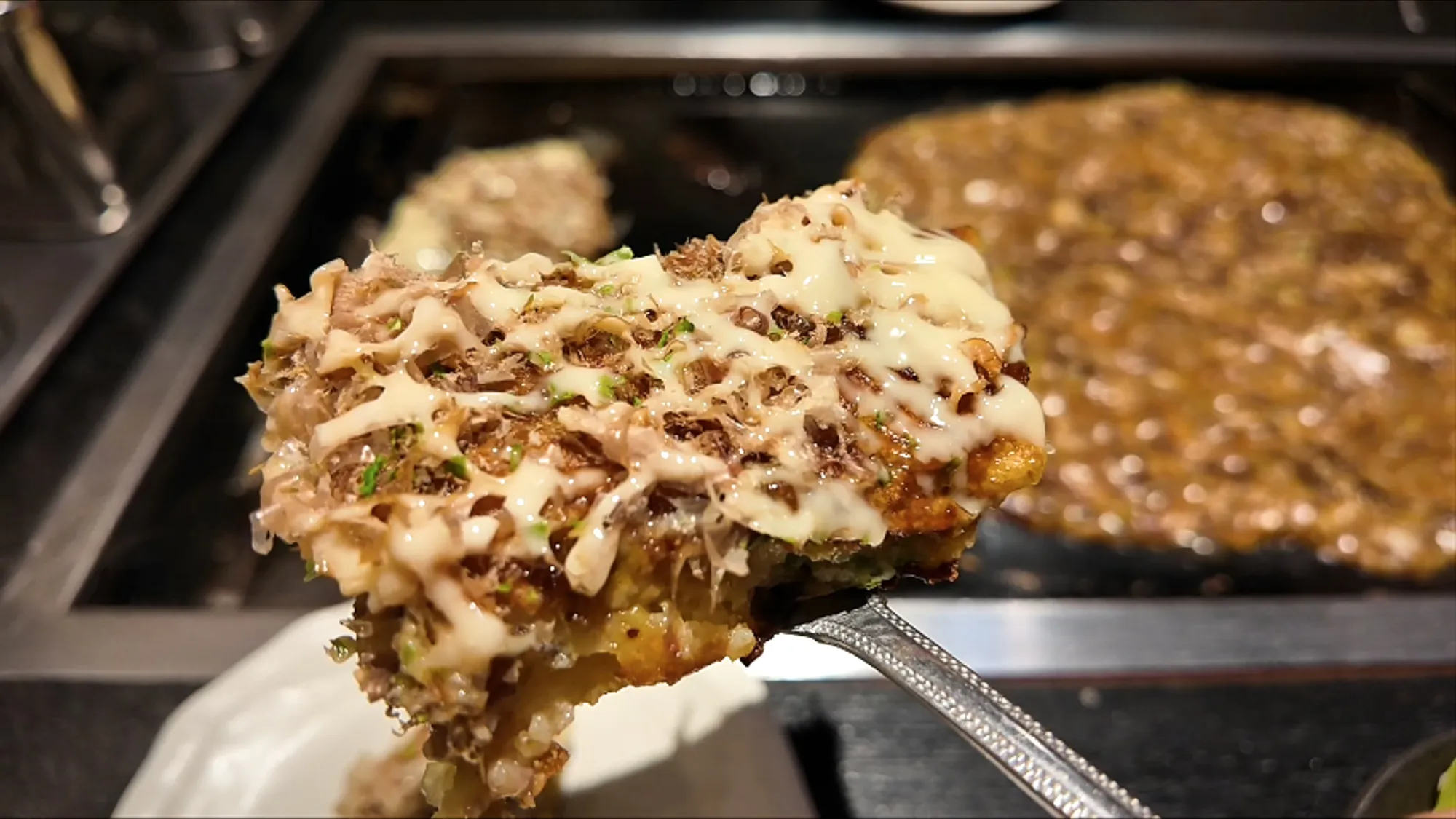 A bite of okonomiyaki with mayonnaise and bonito flakes held up on a small metal spatula over the griddle at Osakaya, Tokyo.