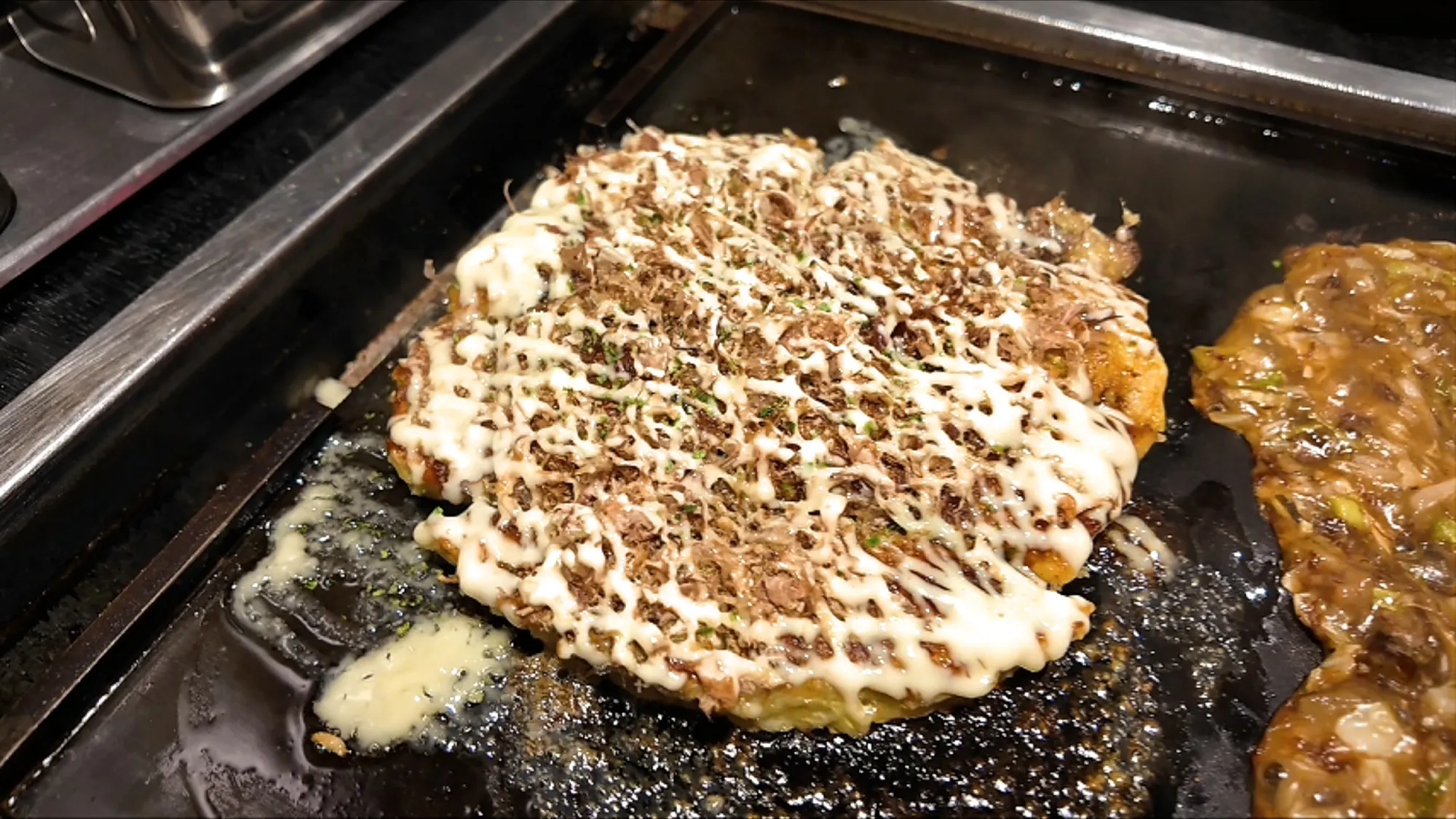 An okonomiyaki covered in mayonnaise and bonito flakes on a teppan griddle at Osakaya in Shinjuku, Tokyo, with monjayaki cooking beside it.