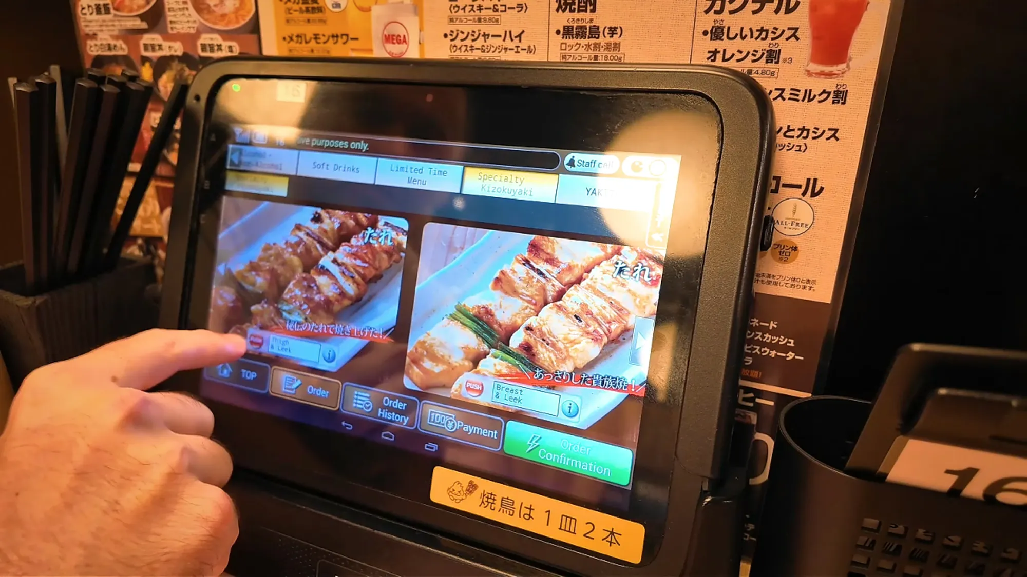 A finger tapping a photo of grilled chicken skewers on the ordering tablet at a Torikizoku table in Tokyo.