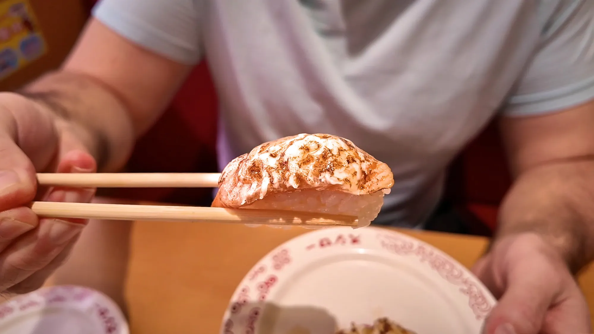 Chopsticks lifting a piece of seared nigiri with a creamy topping over a Sushiro plate in Tokyo.