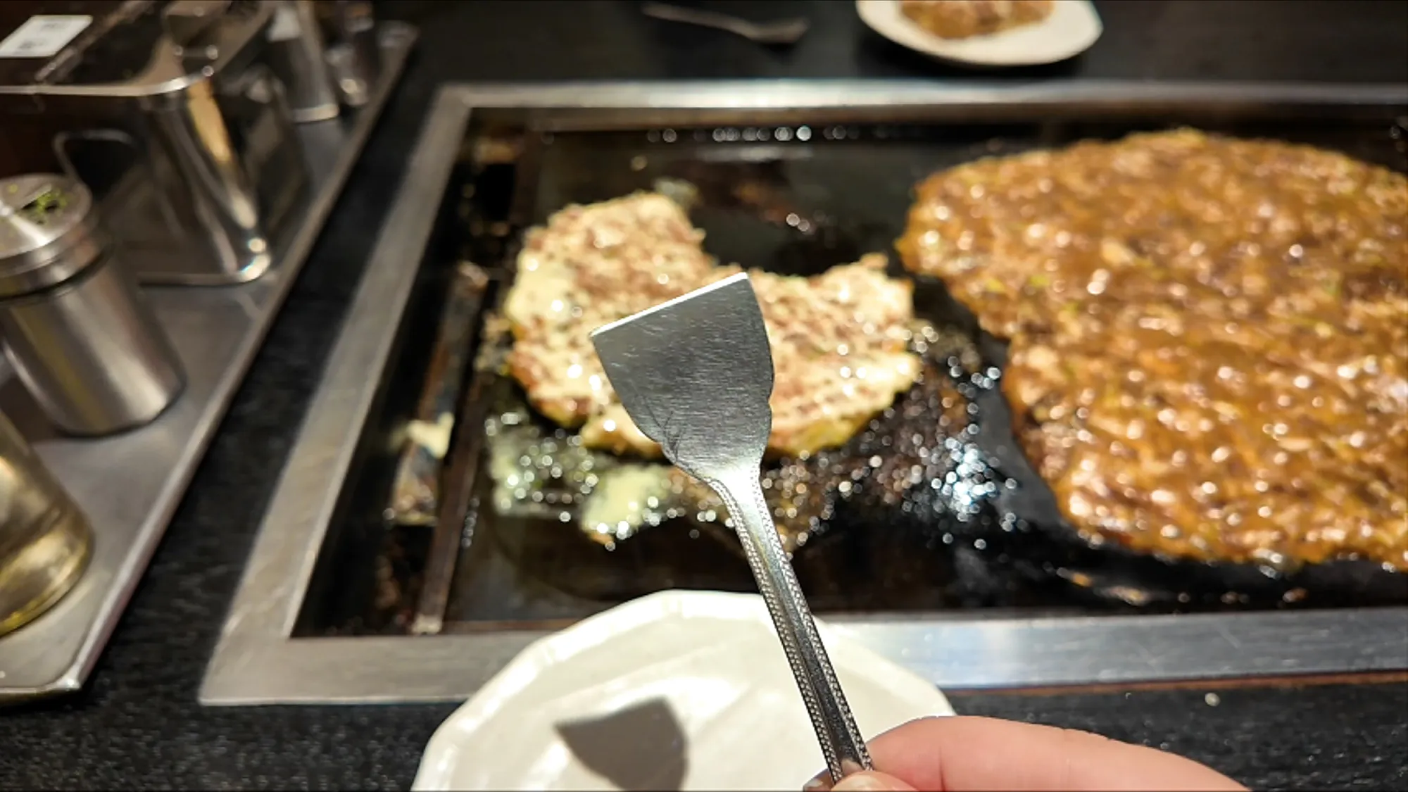 A small metal spatula lifting monjayaki off the teppan griddle at Osakaya, Tokyo, with okonomiyaki and a plate in the background.
