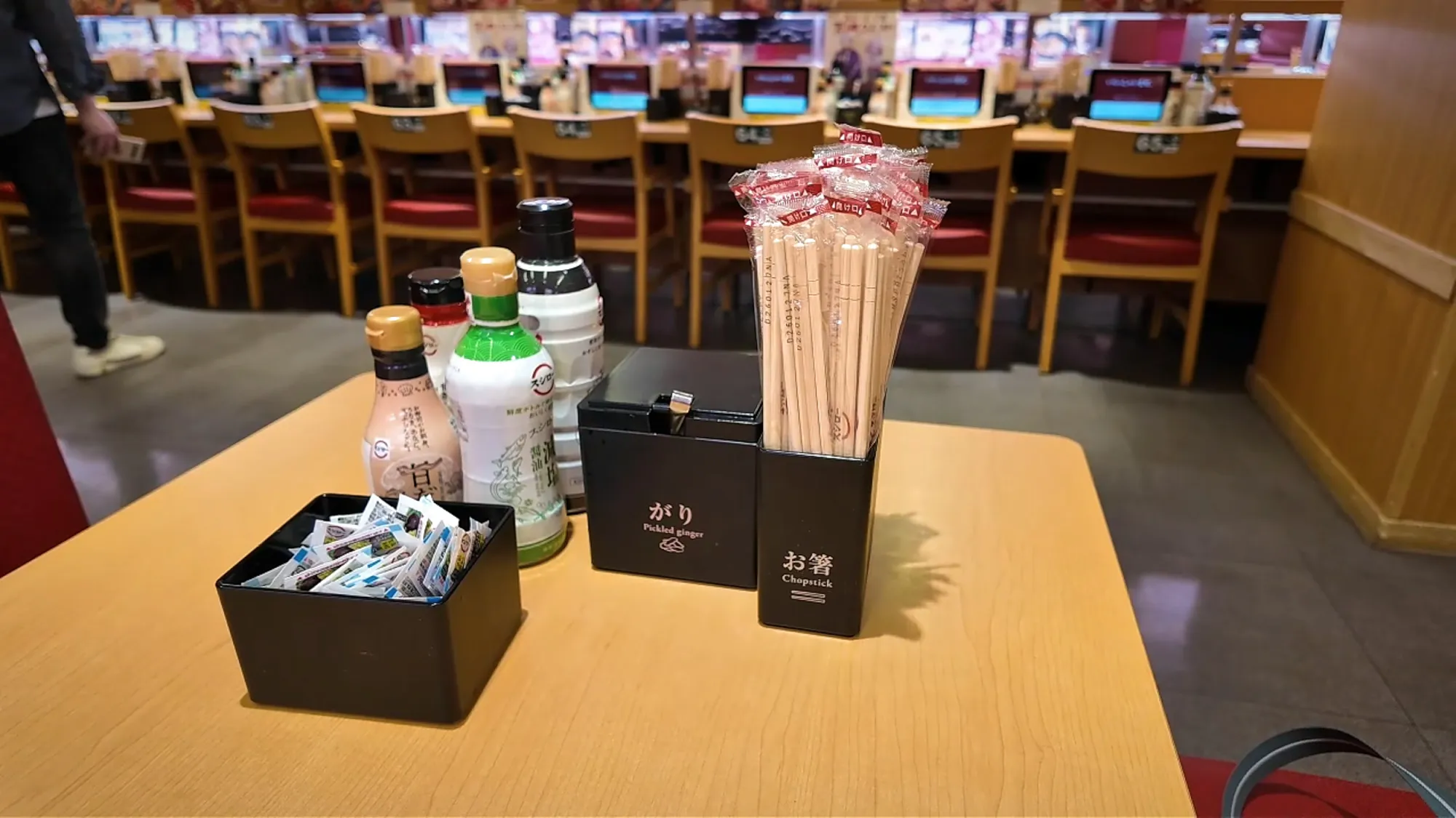 Pickled ginger, chopsticks, and soy sauce bottles waiting on a table at Sushiro in Tokyo, with the conveyor belt counter behind.