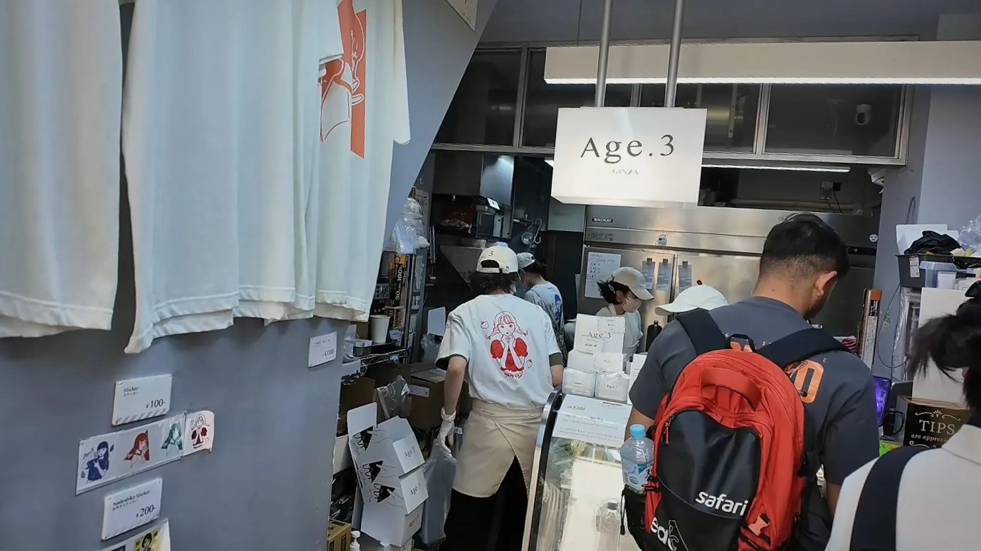 Customers ordering at the small counter inside Age.3 Ginza, with Age.3 T-shirts hanging on the wall and the Age.3 sign above.