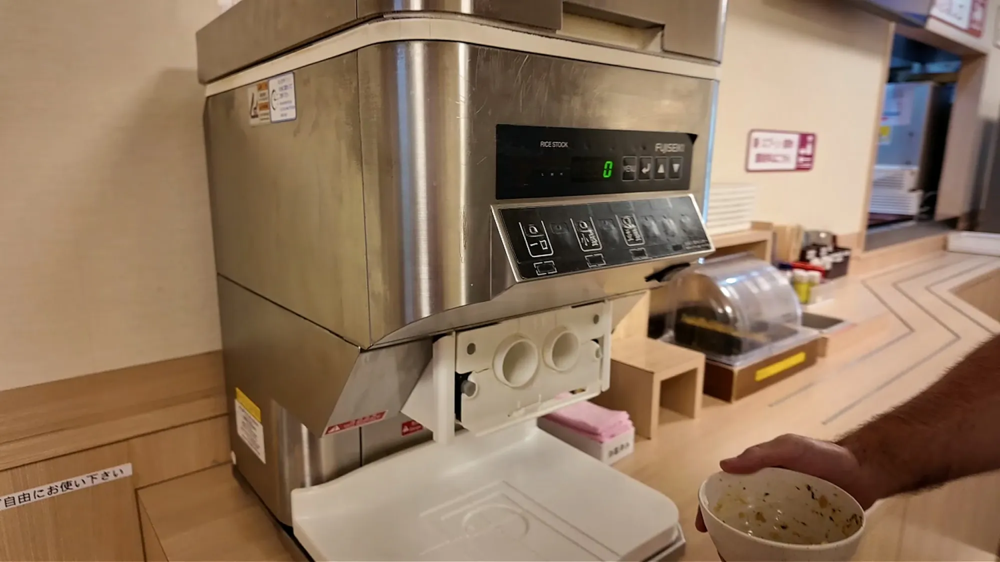 A hand holding an empty rice bowl next to the automatic rice refill machine at Yayoiken in Tokyo.