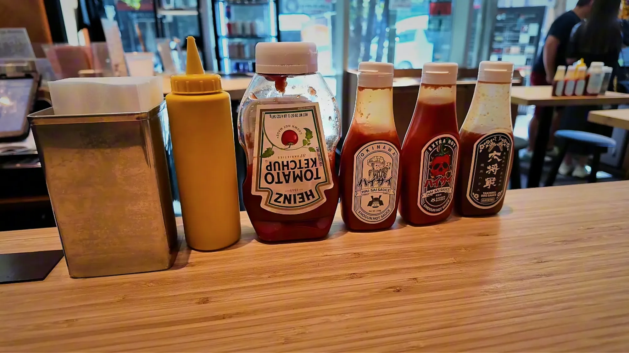 A row of table condiments at Shogun Burger in Shinjuku: a napkin box, mustard, ketchup, and three house sauces.