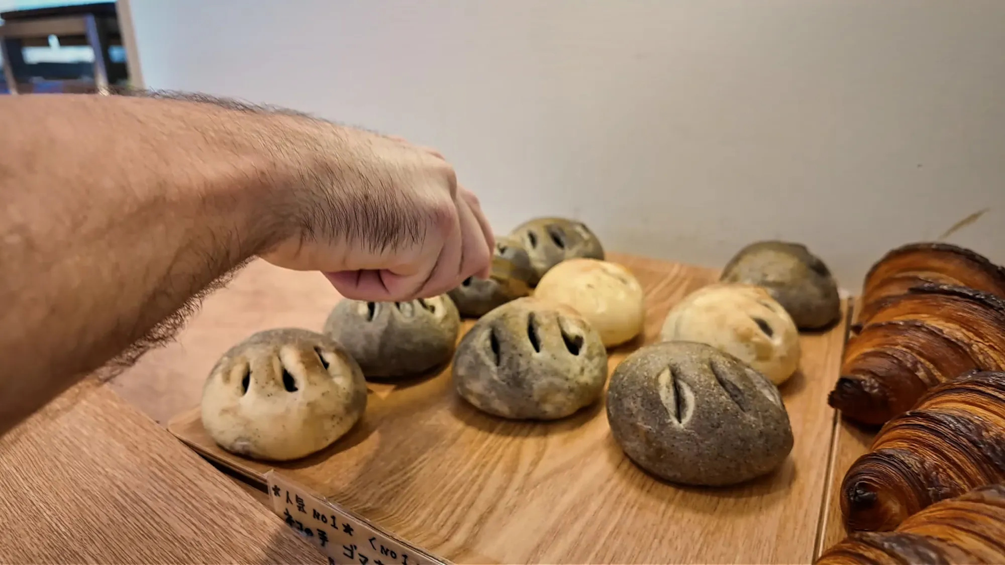 Cream reaching for a tray of round grey black sesame buns with scored tops next to croissants on a wooden shelf inside Donsuke Bakery