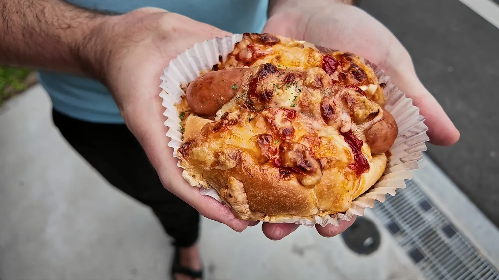 A savory Japanese bakery bun topped with a sausage, browned melted cheese, and red sauce, in a paper liner