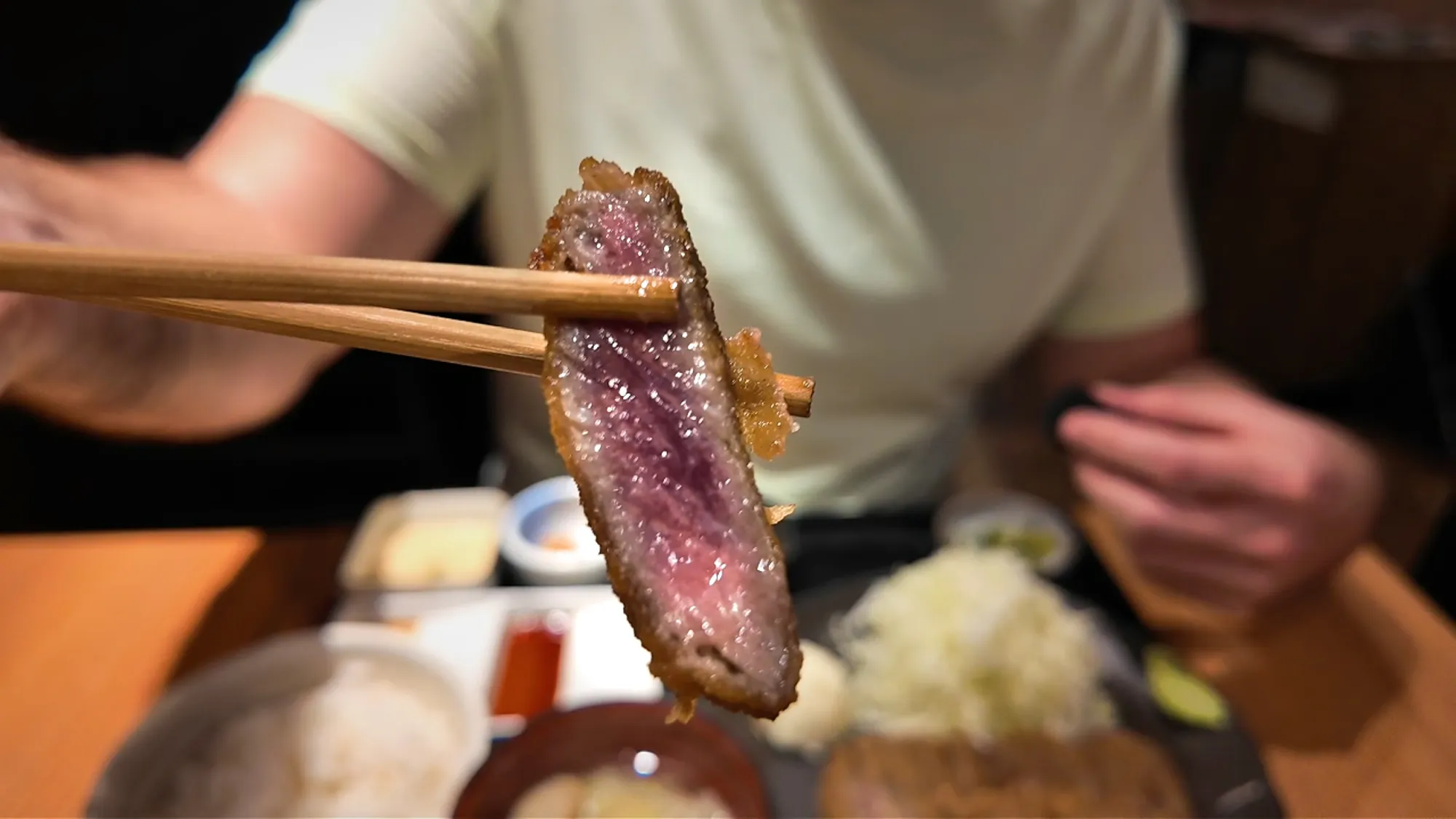 A slice of gyukatsu held up with chopsticks, showing a crisp brown crust and a deep red rare center.
