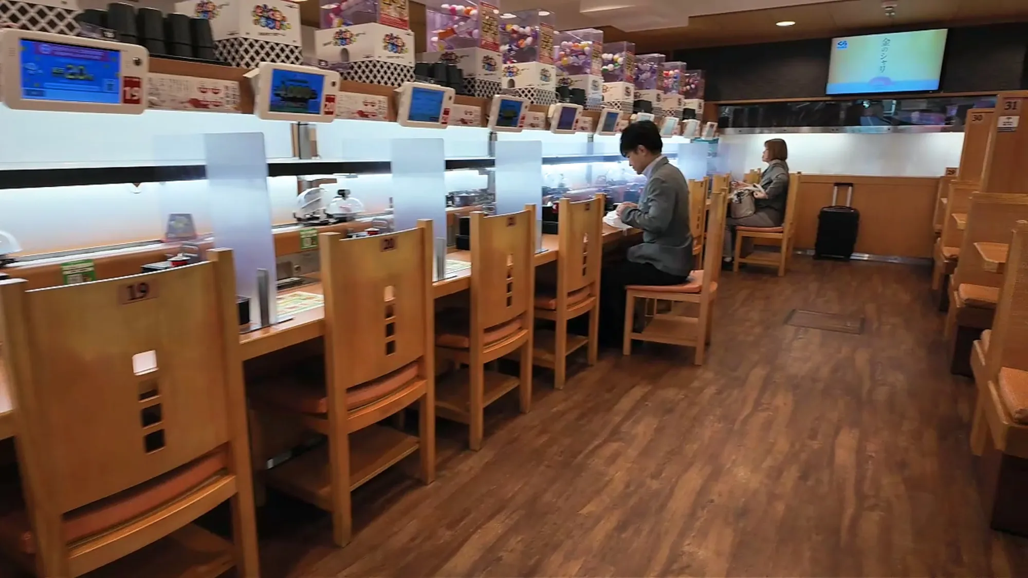 A diner eating alone at a long conveyor belt sushi counter in Tokyo, with rows of single seats and dividers along the belt.