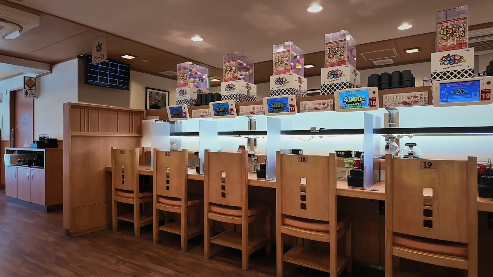 A long row of single wooden seats along a conveyor belt sushi counter in Tokyo, each with its own ordering screen and a divider between seats.