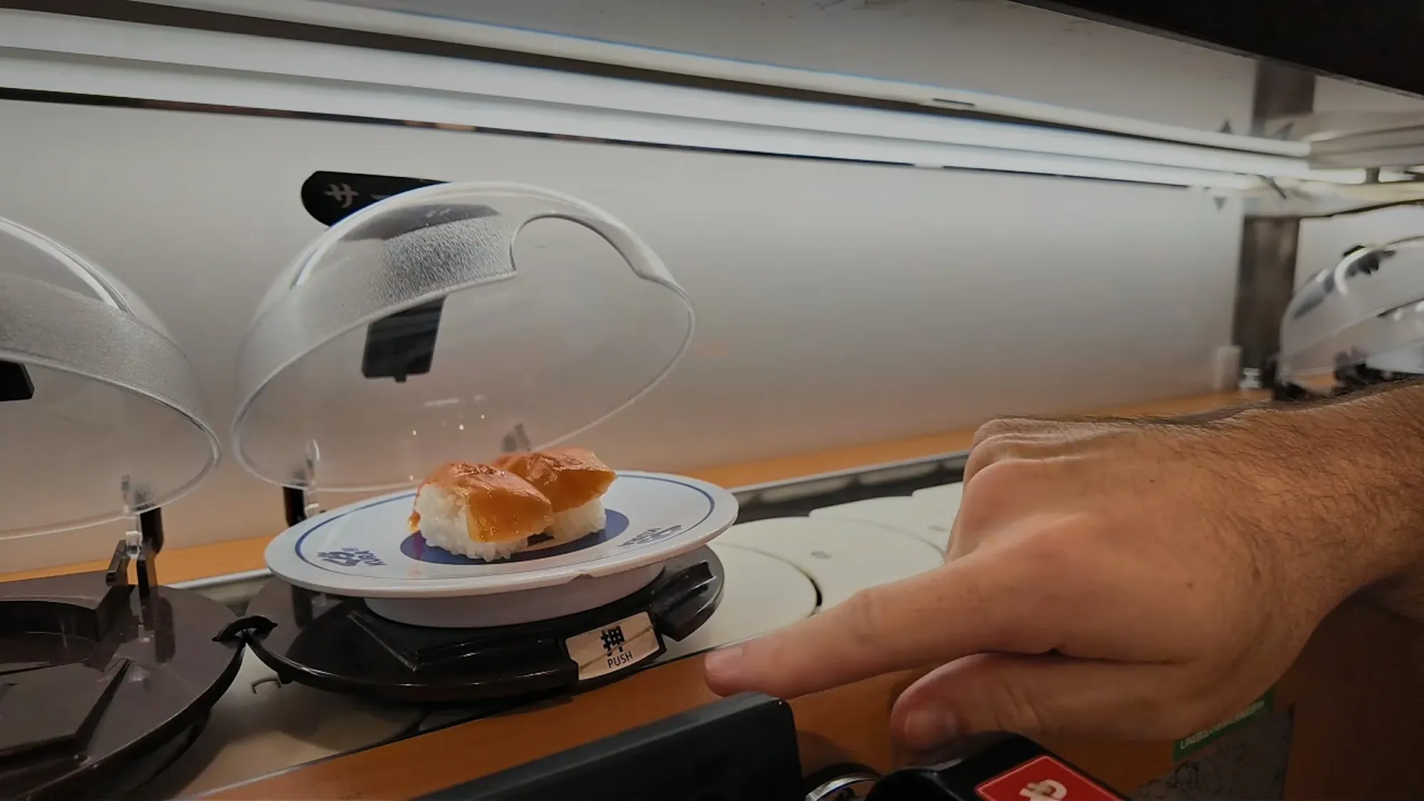 A hand pressing the button that opens the clear dome cover over a plate of sushi on the Kura Sushi conveyor belt.