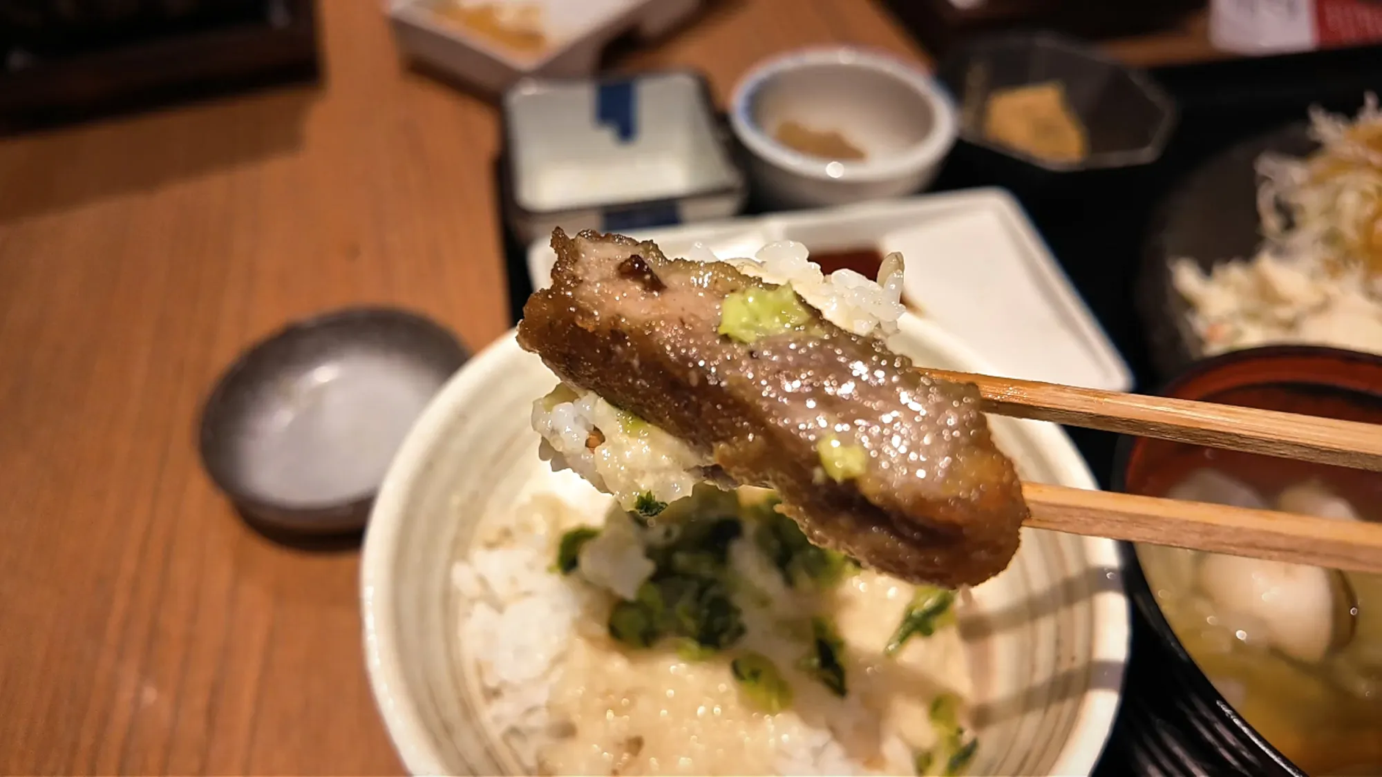 A seared slice of gyukatsu with wasabi held over a bowl of barley rice topped with grated yam and green pickles.
