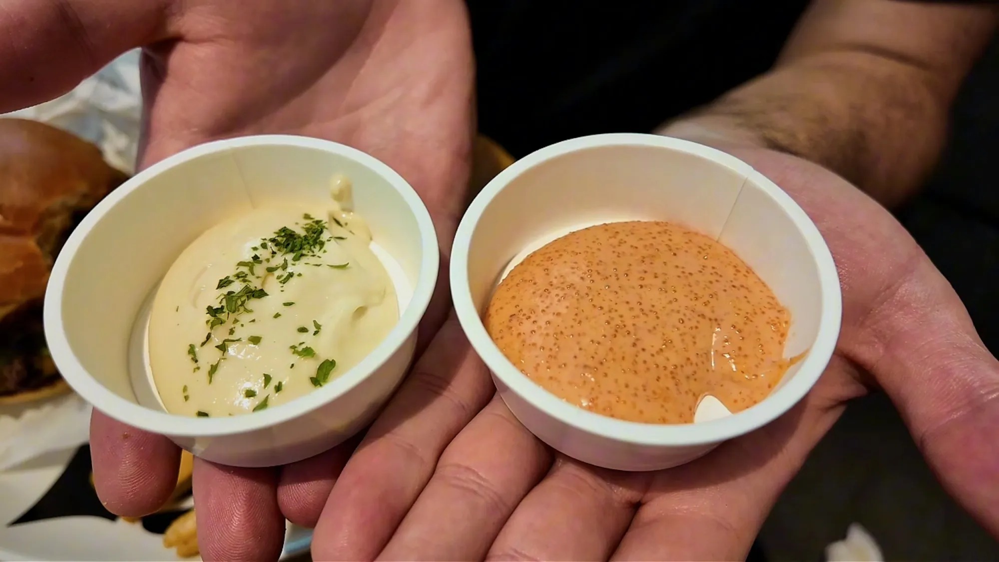 A hand holding two small cups of dipping sauce at Shogun Burger: creamy lemon garlic sauce with herbs and orange mentaiko sauce.