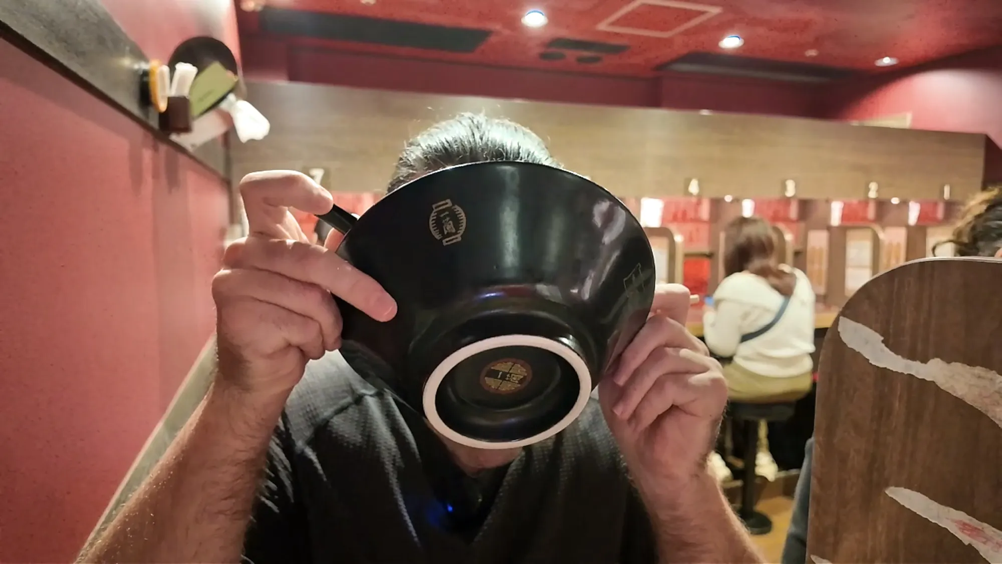 Cream lifting an Ichiran bowl to drink the last of the broth, with a row of red solo booths behind him.