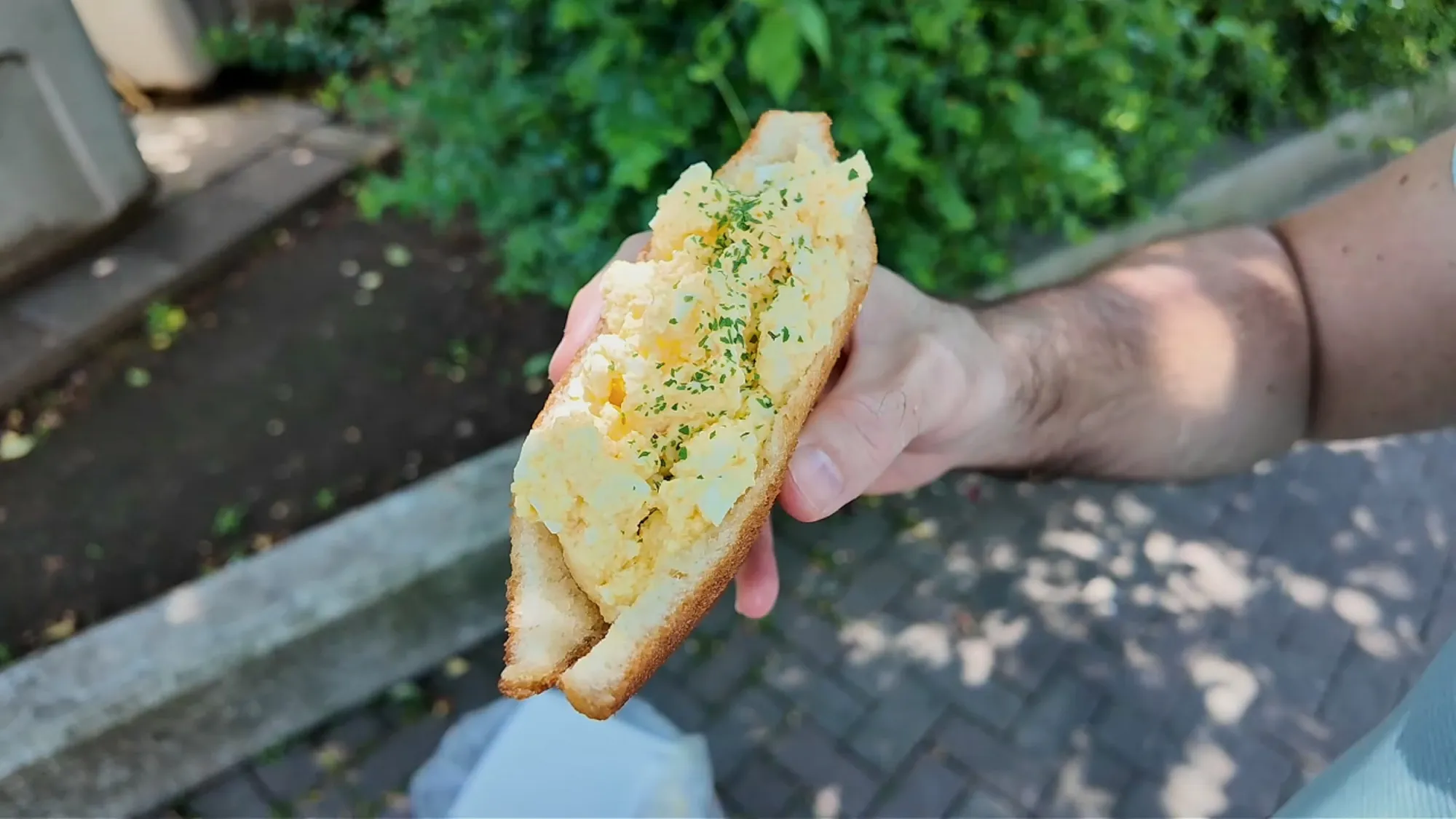 A deep-fried sandwich overstuffed with egg salad and sprinkled with parsley, held up on a shady sidewalk near Age.3 Ginza.
