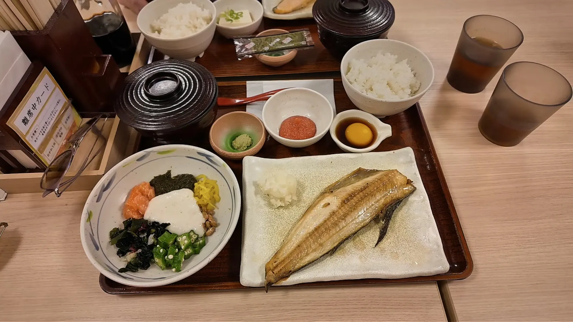 A Japanese breakfast in Tokyo at Yayoiken, a tray with grilled shima hokke fish, a bowl of rice, miso soup, mentaiko, a raw egg in soy sauce, and a bowl of natto, okra, and grated yam.