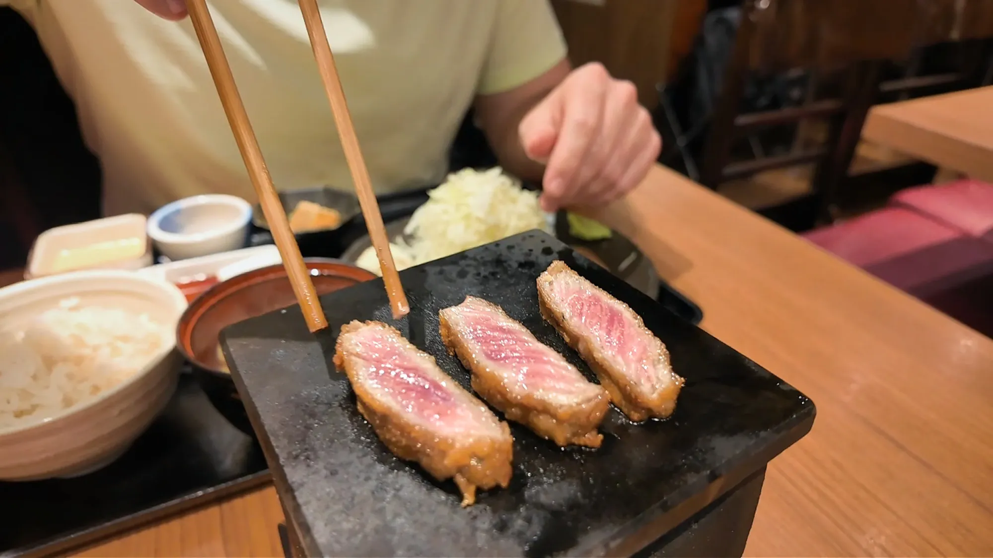 Chopsticks laying rare slices of breaded beef cutlet on a hot stone at Gyukatsu Motomura in Tokyo.