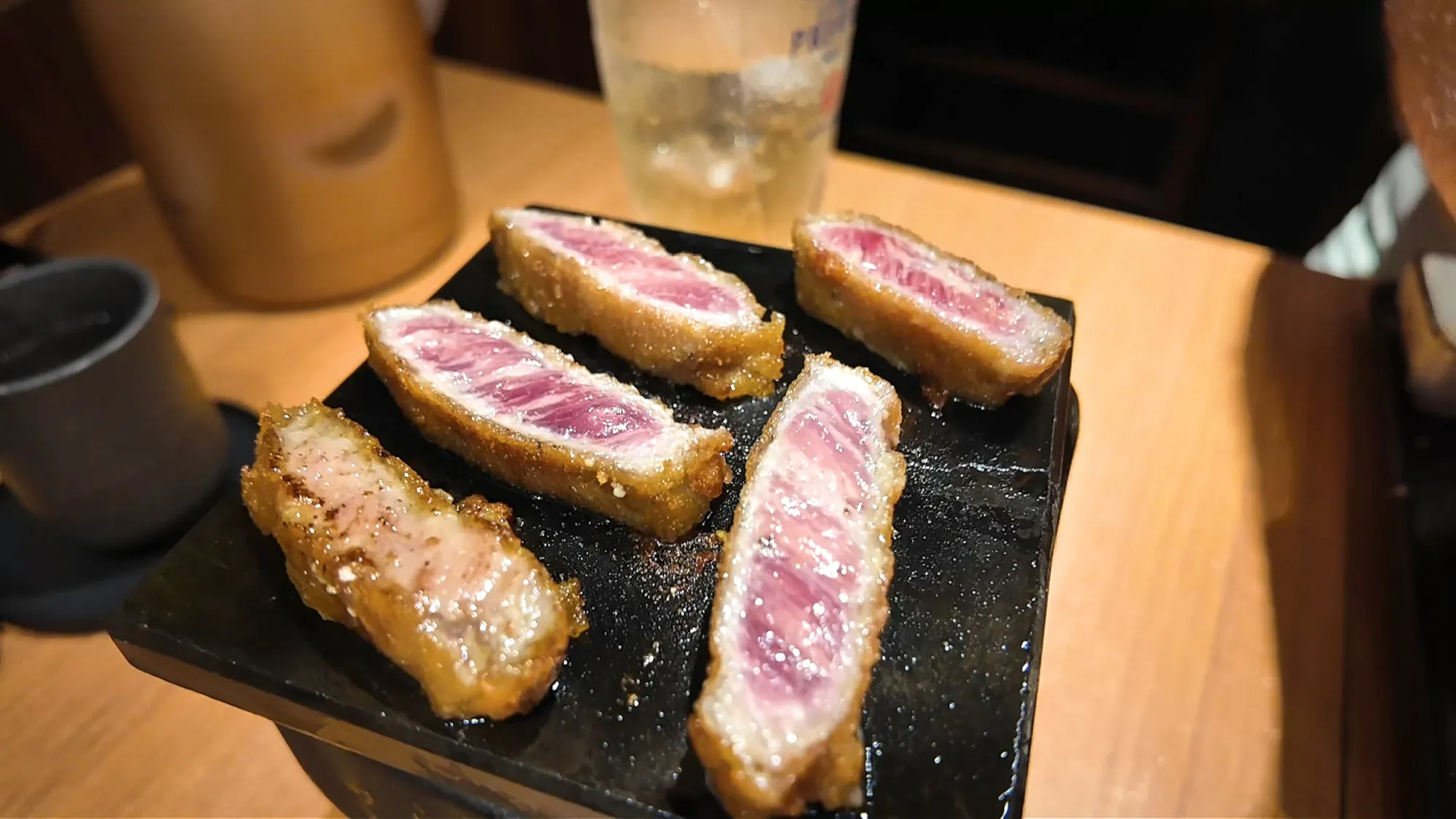 Rare slices of breaded gyukatsu beef cutlet with pink centers on a black hot stone at Gyukatsu Motomura in Tokyo.