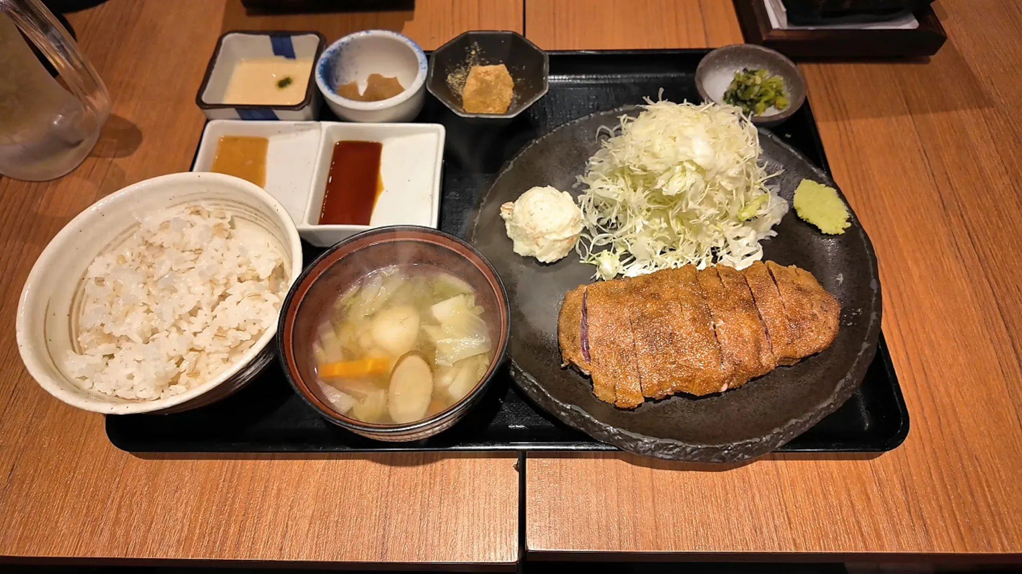 A Gyukatsu Motomura set on a black tray with barley rice, soup, a sliced beef cutlet with shredded cabbage, potato salad and wasabi, two sauces, and three small side dishes.