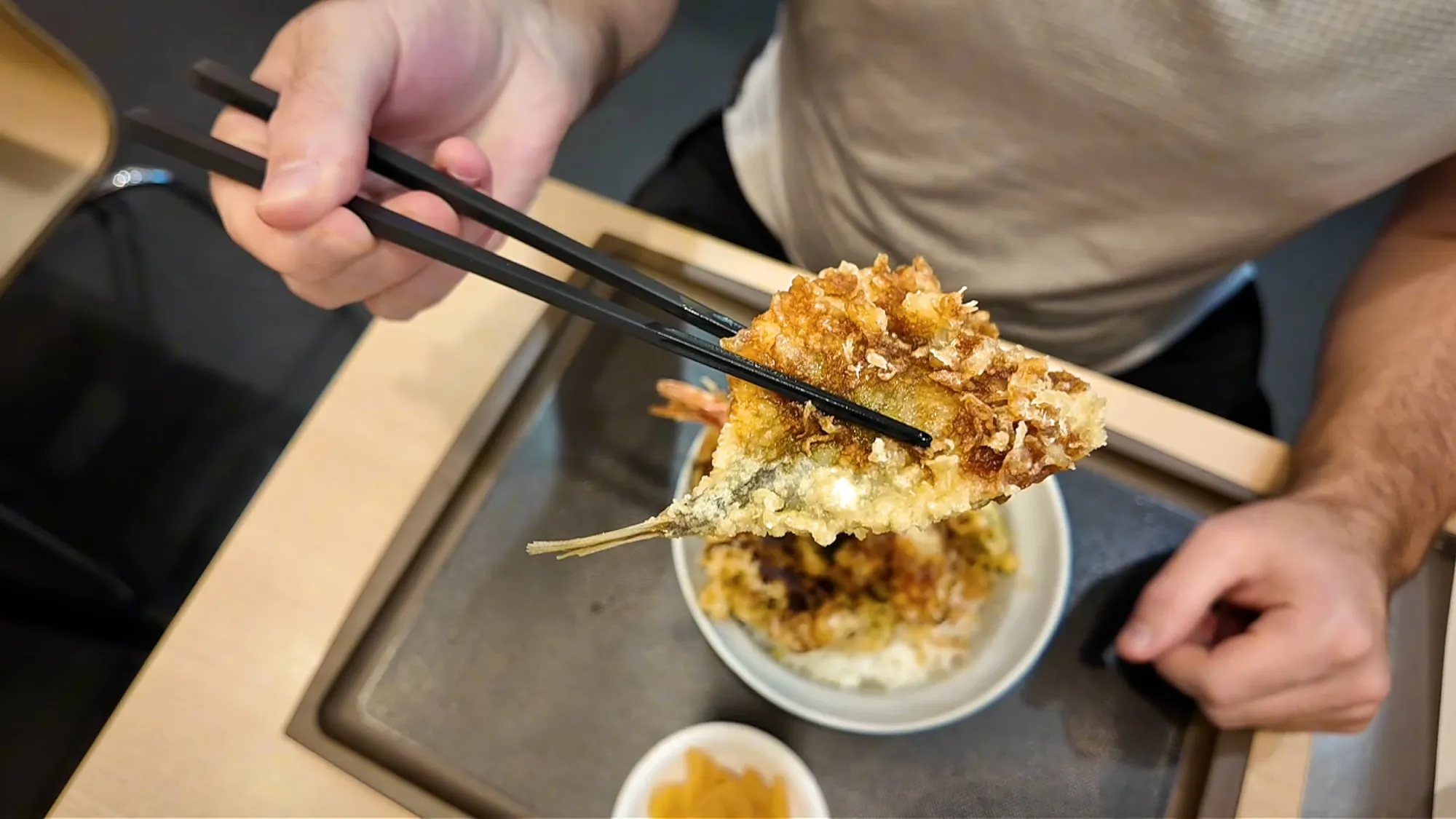 Cream holding up a piece of horse mackerel tempura with chopsticks over the Sea Breeze Tendon at Tenya.