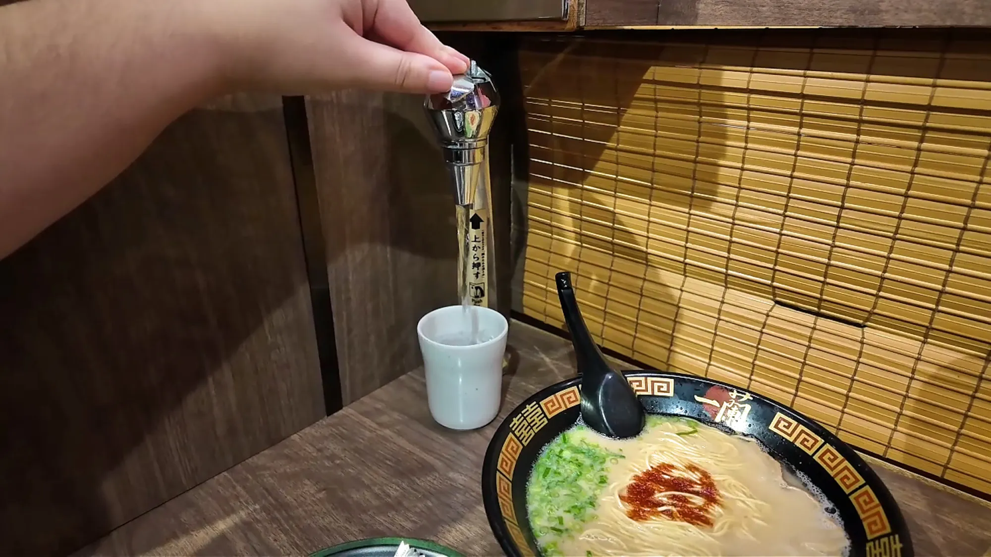 Filling a cup from the private water tap in an Ichiran solo booth, with a bowl of tonkotsu ramen on the counter.