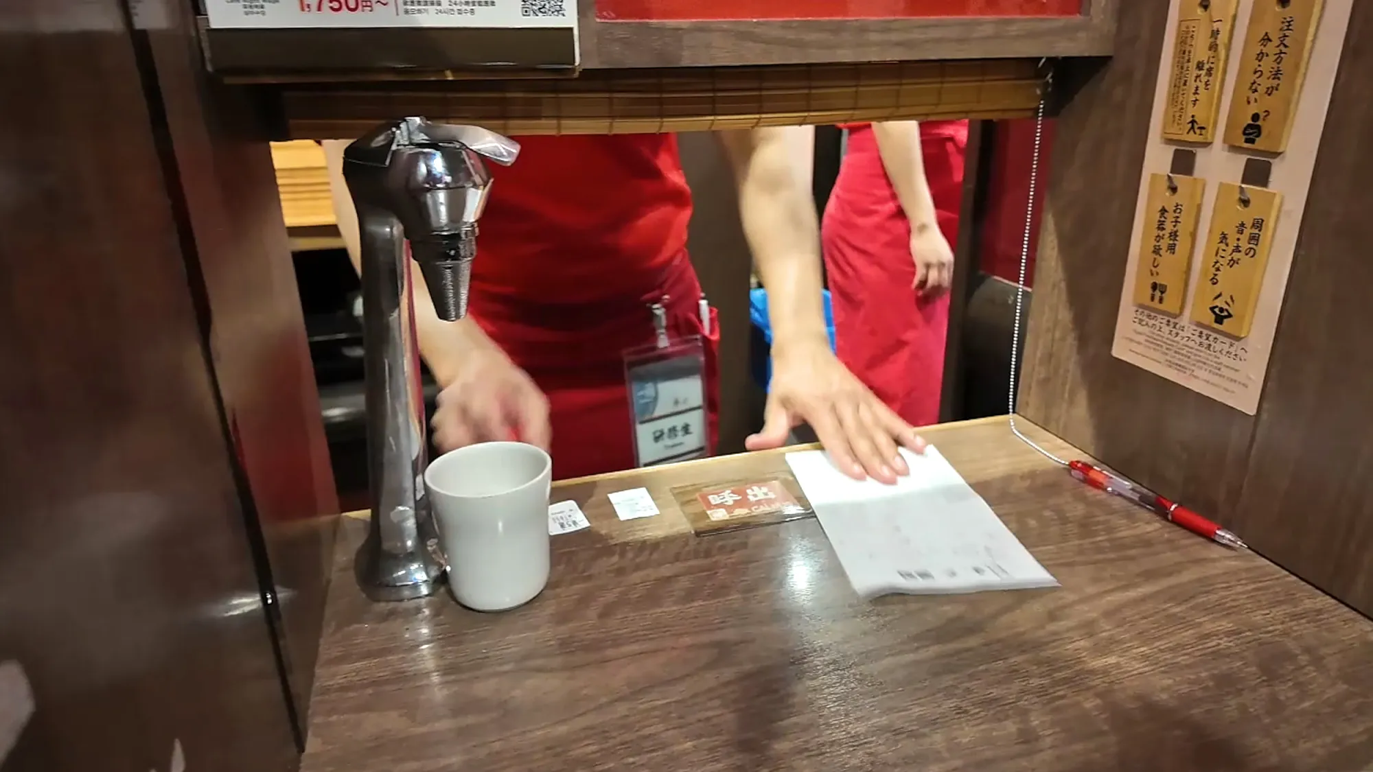 An Ichiran staff member in red reaching through the booth window to collect the order sheet, beside the booth's water tap and a cup.