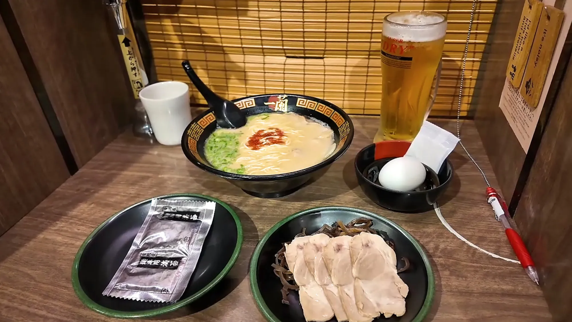 A bowl of Ichiran tonkotsu ramen with red sauce and green onion in a wooden solo booth, with a draft beer, a salted egg, sliced chashu pork, and wrapped seaweed in Tokyo.